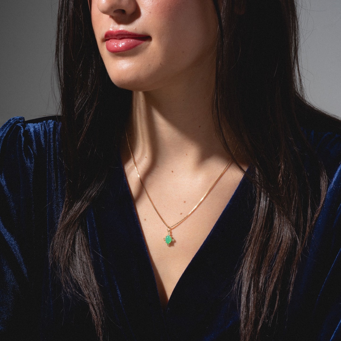 A woman with long dark hair wears a dark blue velvet top and a gold One of a Kind 14k Chrysoprase and Diamond Charm necklace, shown from the shoulders up.