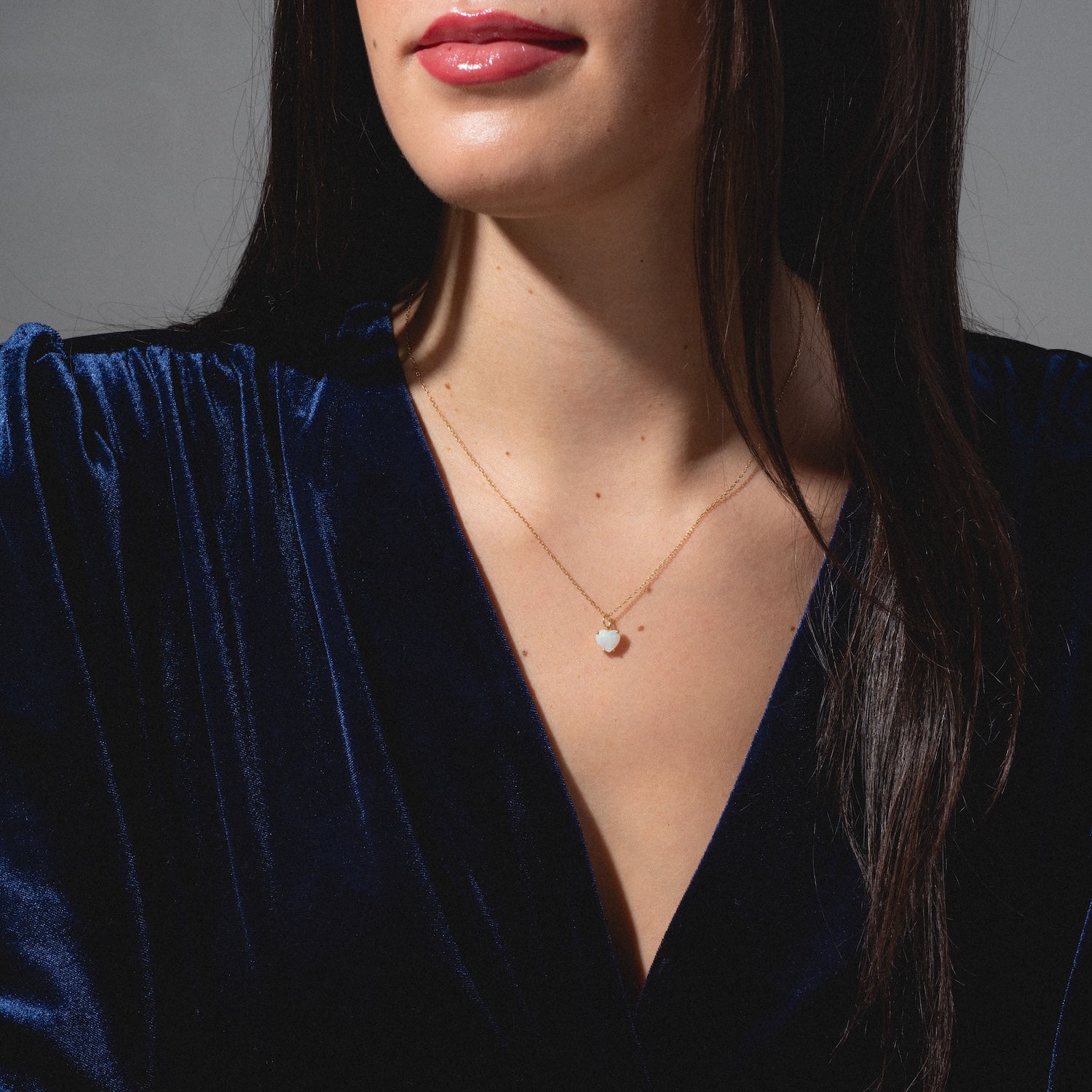 A woman wears a deep blue velvet dress and the One of a Kind 14k Opal Heart Charm necklace, shown from the shoulders up with her face partially visible.