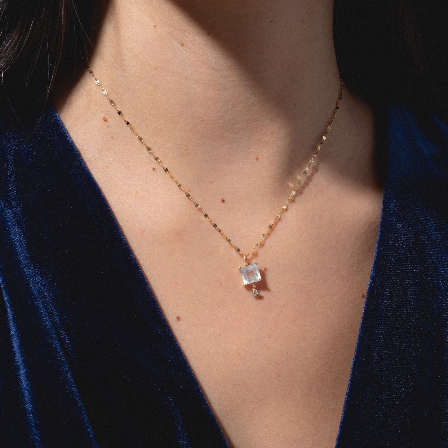 A close-up of a person wearing the One of a Kind 14k Princess Cut Rainbow Moonstone with Diamond Dangle Charm necklace, styled with a deep blue velvet top.