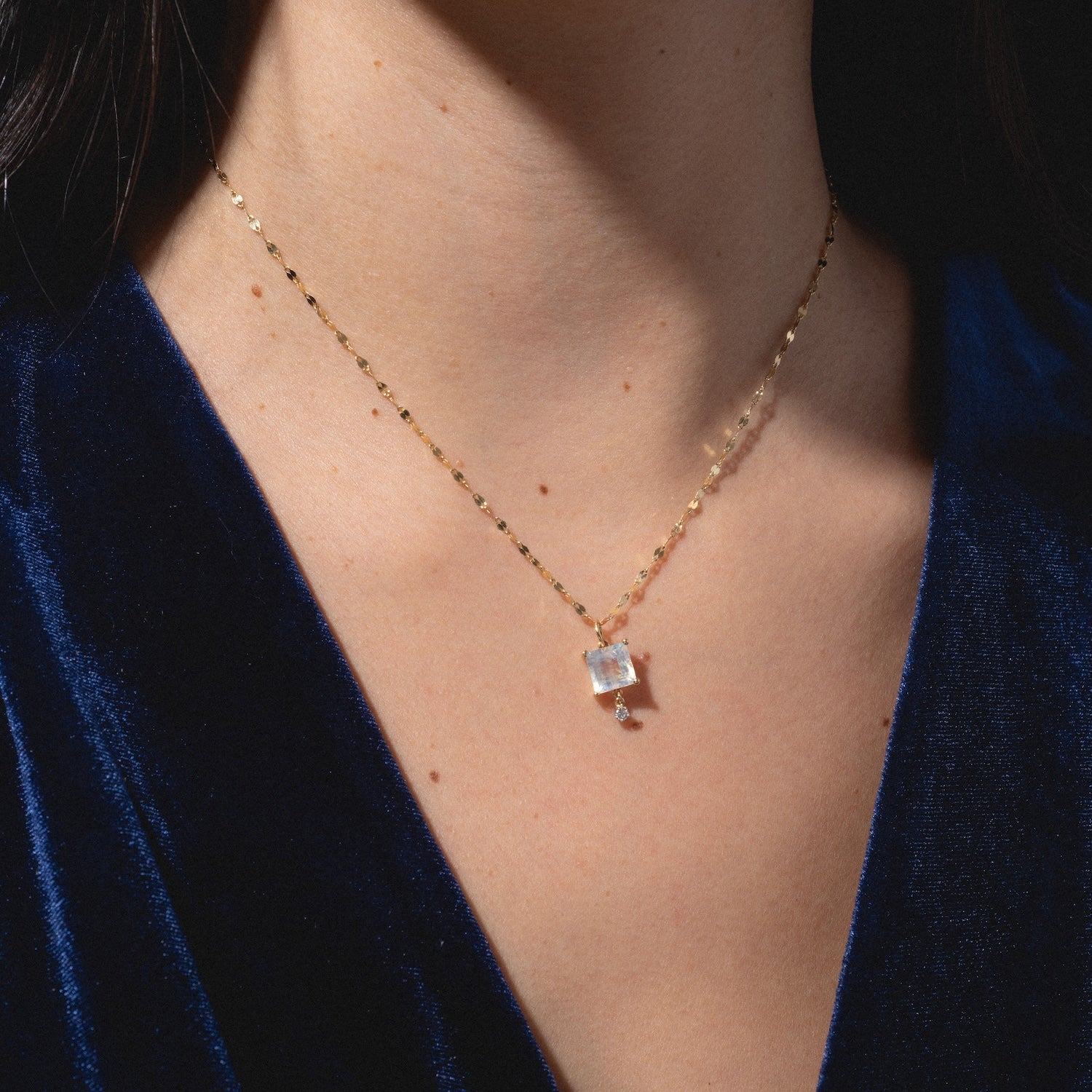 A close-up of a person wearing the One of a Kind 14k Princess Cut Rainbow Moonstone with Diamond Dangle Charm necklace, styled with a deep blue velvet top.