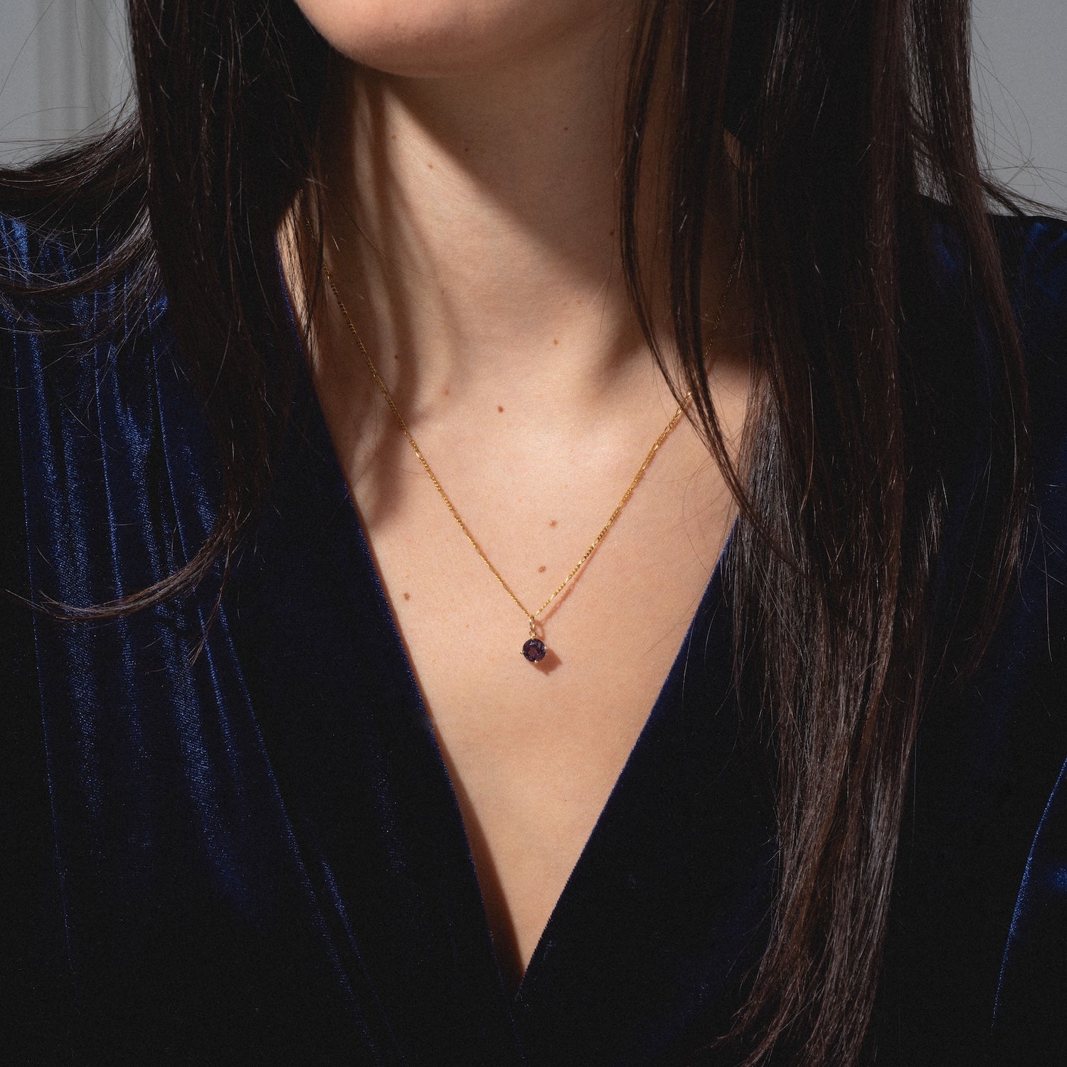 A woman with long dark hair wears a deep blue velvet top and a delicate gold necklace featuring the One of a Kind 14k Round Lab Alexandrite Charm. The focus is on her neck and chest, with her face partially out of the frame.