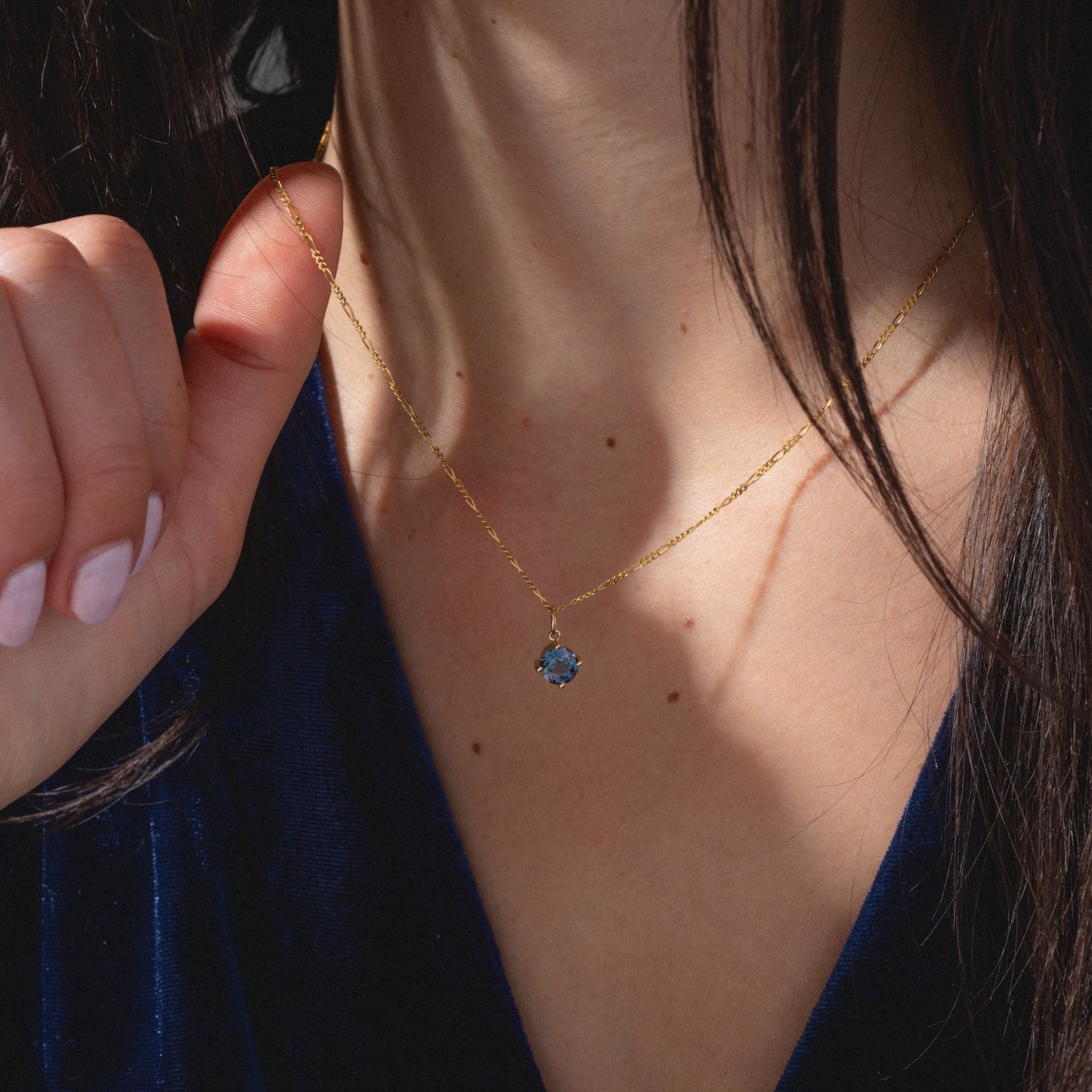 A woman in a deep blue velvet top holds the One of a Kind 14k Round Lab Alexandrite Charm, a delicate gold chain necklace with a blue gemstone pendant, to her neck as her long brown hair partially obscures her face.