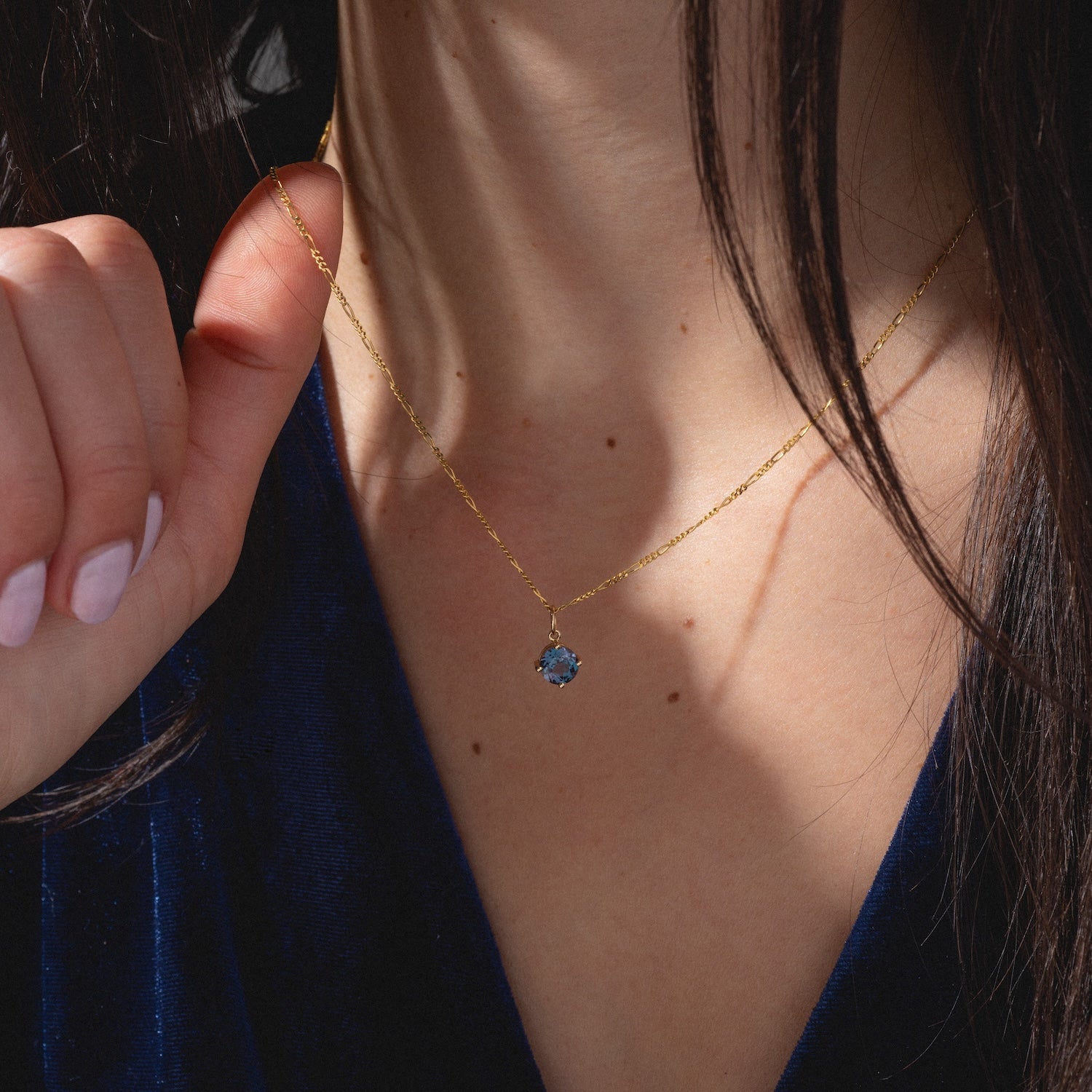 A woman in a deep blue velvet top holds the One of a Kind 14k Round Lab Alexandrite Charm, a delicate gold chain necklace with a blue gemstone pendant, to her neck as her long brown hair partially obscures her face.