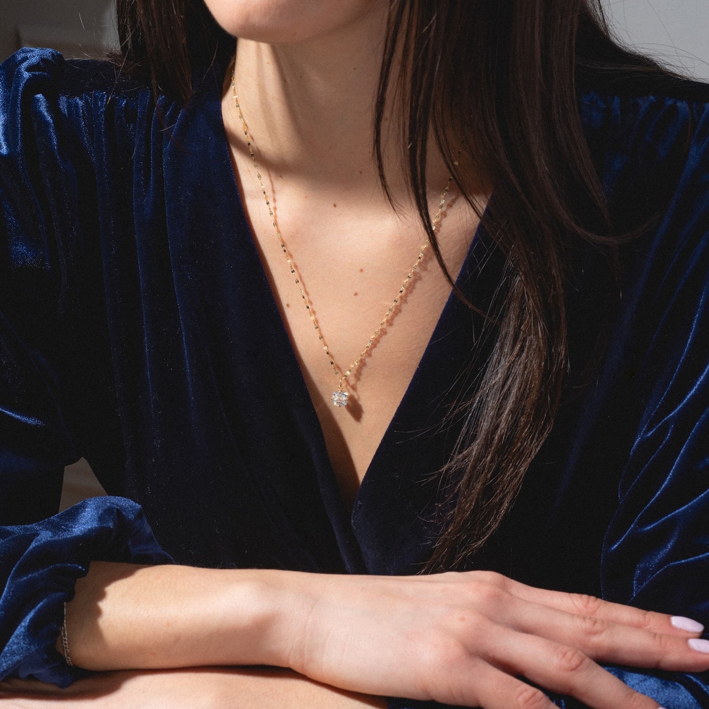 A woman in a deep blue velvet dress wears a One of a Kind 14k Oval Moissanite Charm with hand engraved details, its clear gemstone pendant glinting in the sunlight as she sits at a table with her arms crossed.