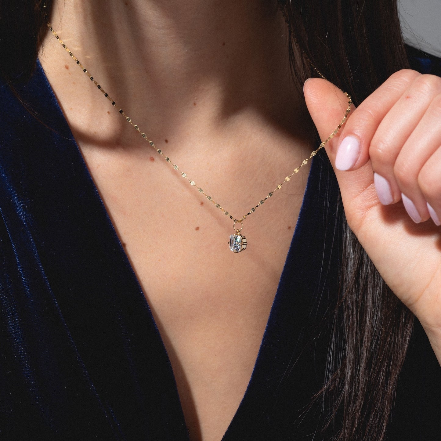 A woman in a dark blue velvet top holds the One of a Kind 14k Oval Moissanite Charm with Hand Engraved Details near her neck, showcasing its sparkling pendant. Her nails are manicured with pale pink polish.