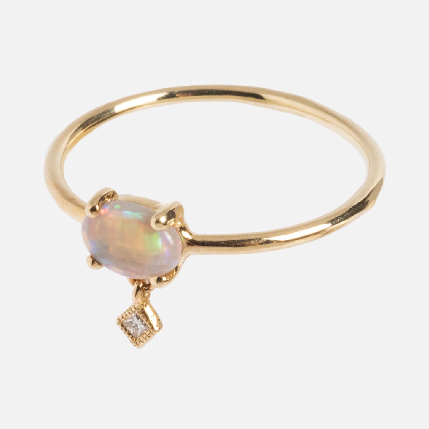 The One of a Kind 14k Sweetest Opal with Diamond Dangle Ring showcases an oval opal in a four-prong setting, highlighted by a delicate dangling square diamond charm.