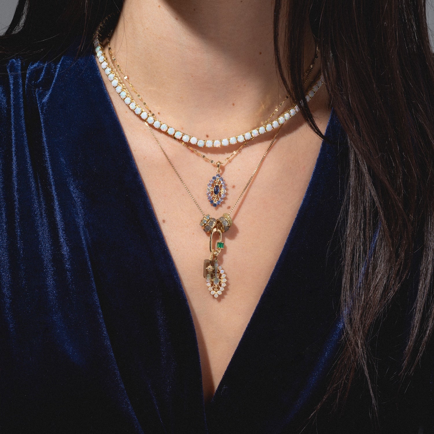 A woman in a dark blue velvet top wears layered gold necklaces, including the One of a Kind 14k Angel Aura and Diamond Charm with gemstones and pearls. Her dark hair partially covers her shoulders.