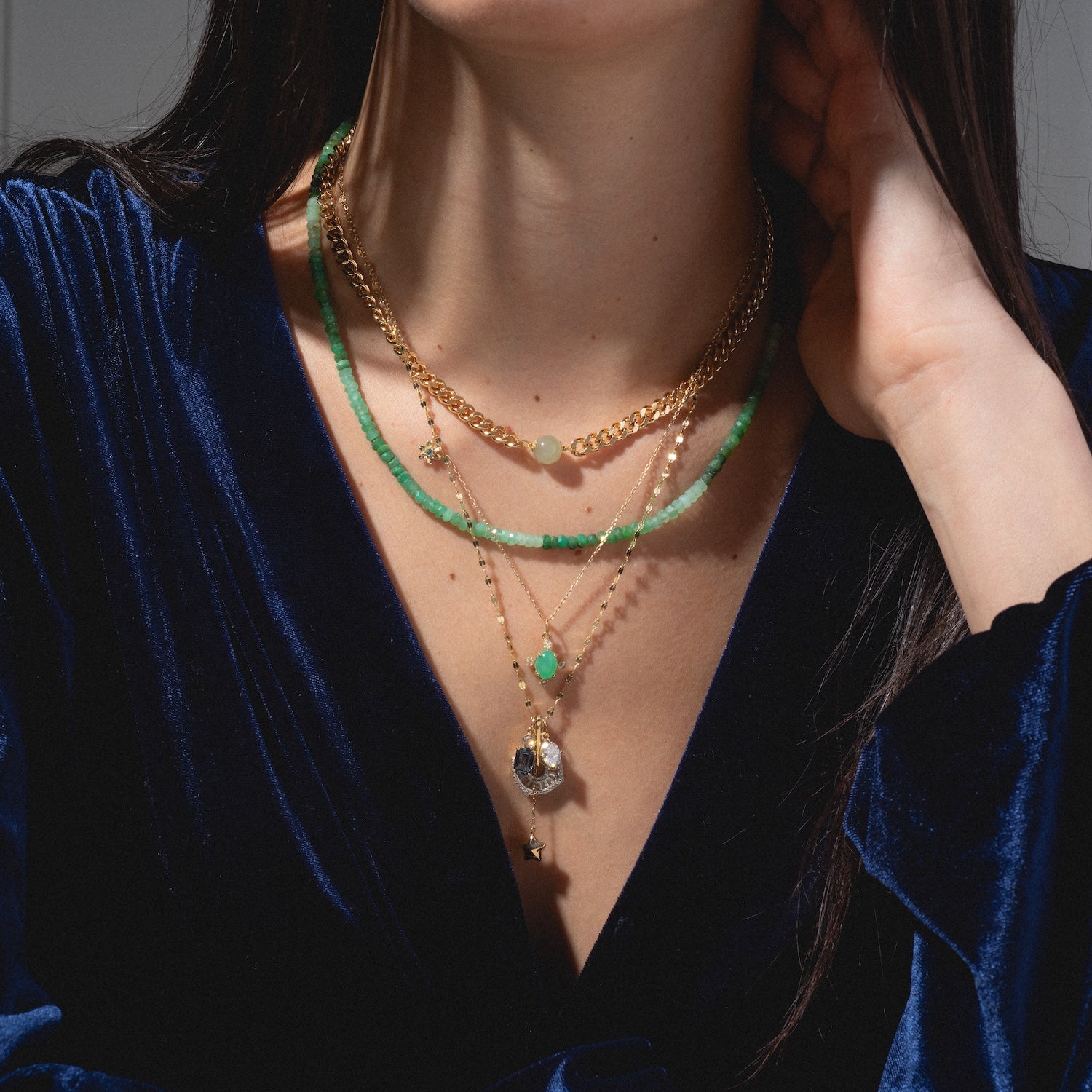 A woman in a deep blue velvet top wears layered necklaces, including green beads, gold chains, and the One of a Kind 14k Chrysoprase and Diamond Charm with stone and star details. She gently touches her neck.