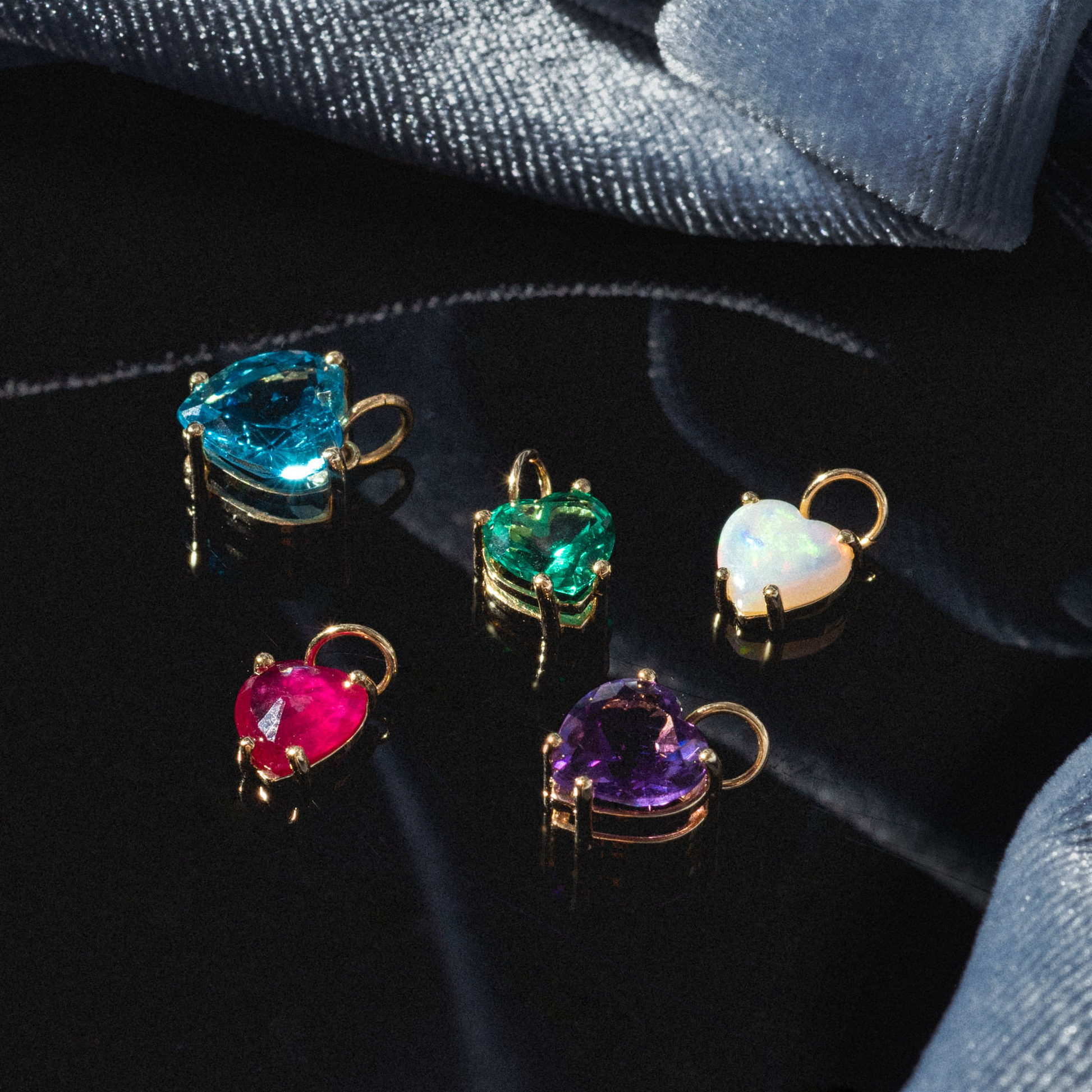 A One of a Kind 14k Opal Heart Charm shines on a dark, glossy surface with blue velvet fabric in the background.