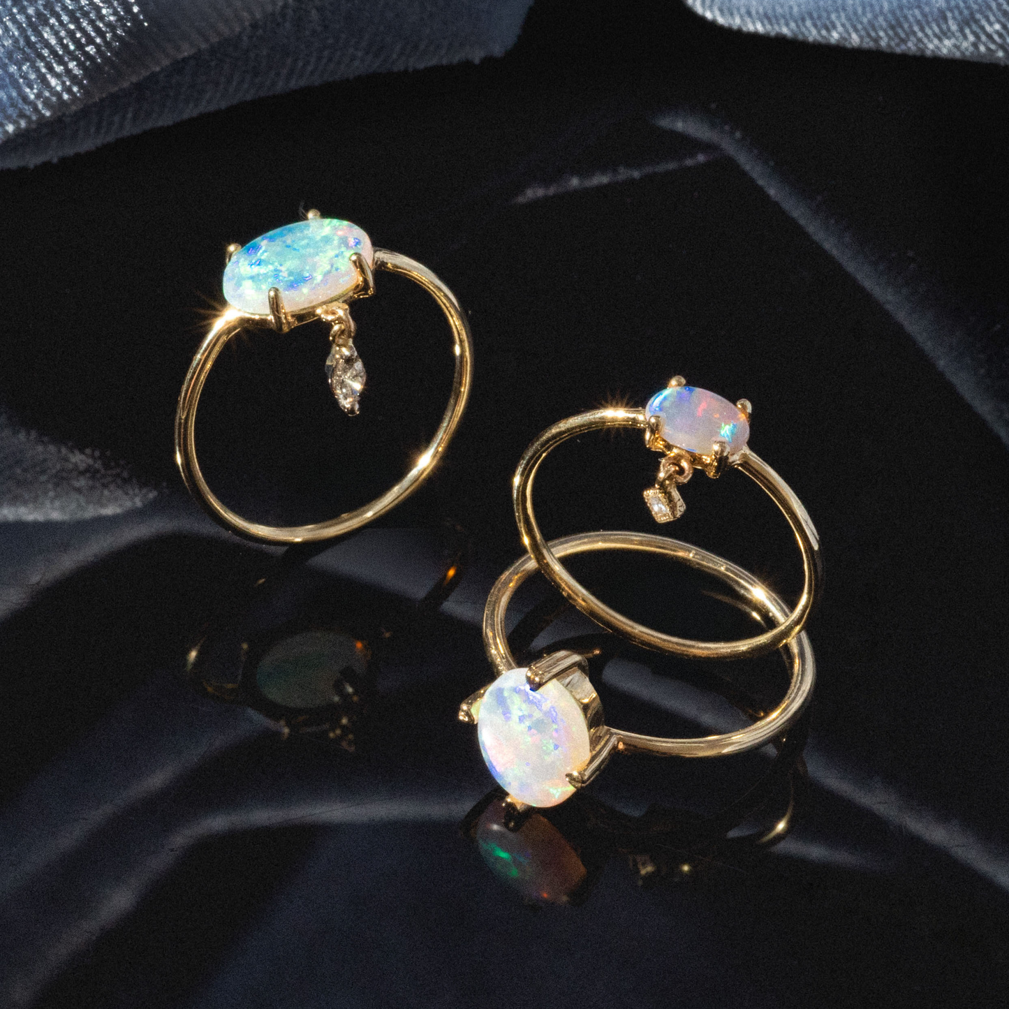 The One of a Kind 14k Heart Prong Opal Ring features an oval opal gemstone with iridescent colors, accented by small dangling diamonds, displayed on a dark reflective surface against soft blue fabric.