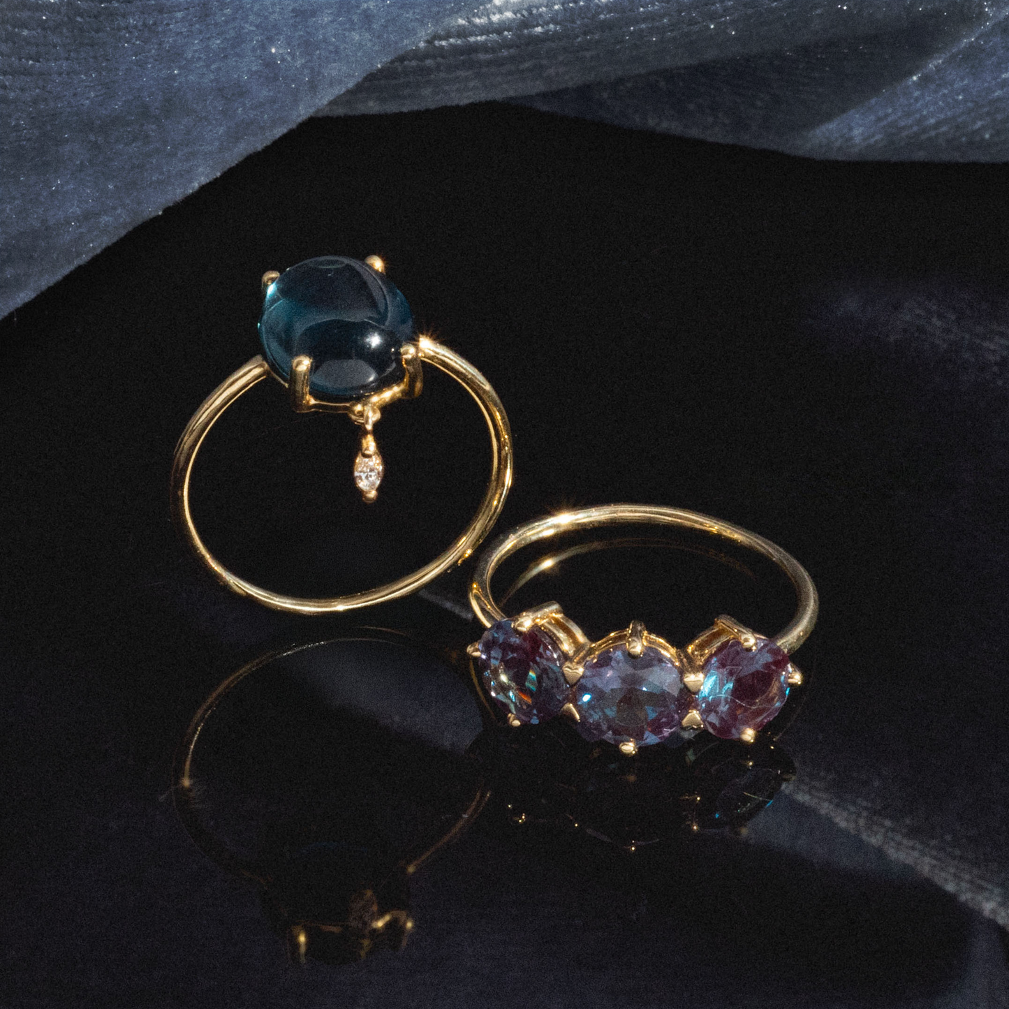Two gold rings rest on a dark surface; one is the One of a Kind 14k London Blue Topaz with Diamond Dangle Ring, featuring an oval blue stone and dangling diamond. The other has three faceted iridescent stones, against draped dark fabric.