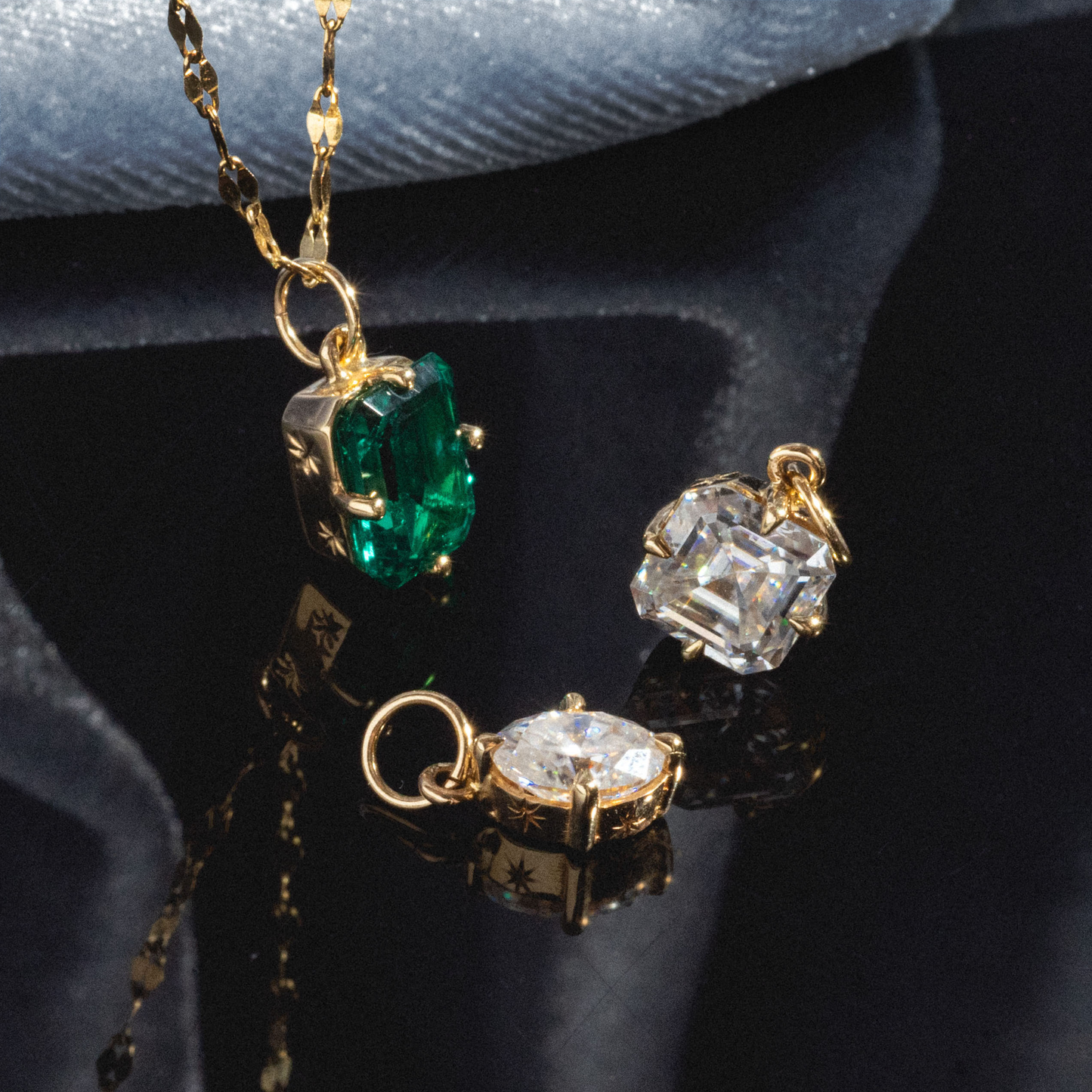 The One of a Kind 14k Lab Emerald Charm with Hand Engraved Details is showcased on a reflective surface beside two gold charms with clear gemstones, all set against a dark, velvety background.