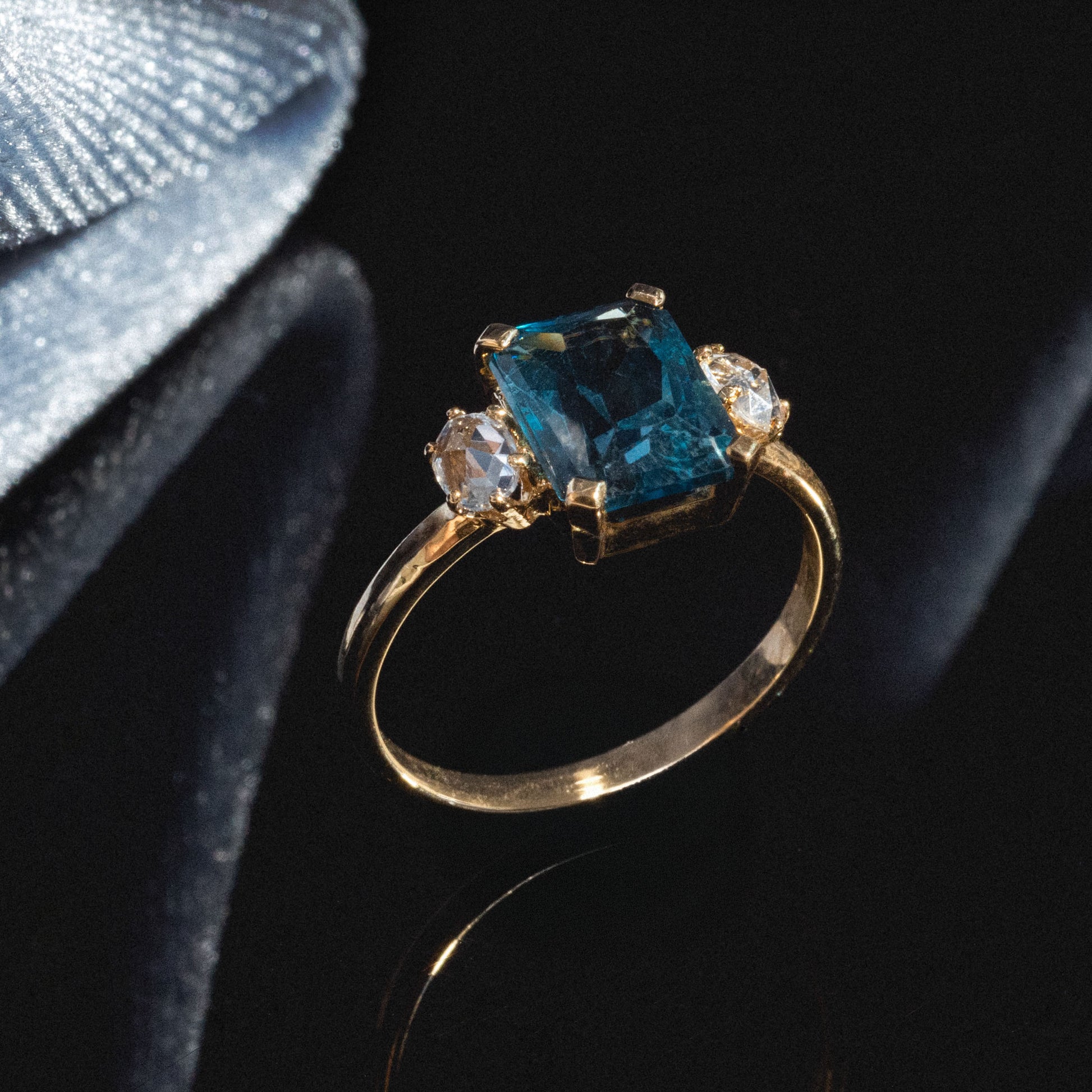 The One of a Kind 14k Vintage Inspired London Blue Topaz and White Sapphire Ring features a large rectangular blue topaz center stone with two white sapphires, displayed on glossy black near sparkly silver fabric.