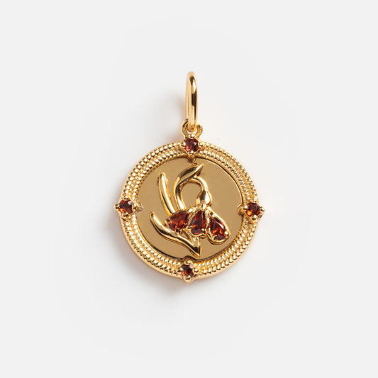 The 2026 January Capsule Vintage Inspired Garnet Coin Charm is a round gold vermeil pendant with a raised orchid center, red gemstone accents, and an ornate border—an exclusive vintage-style piece from Local Eclectic.