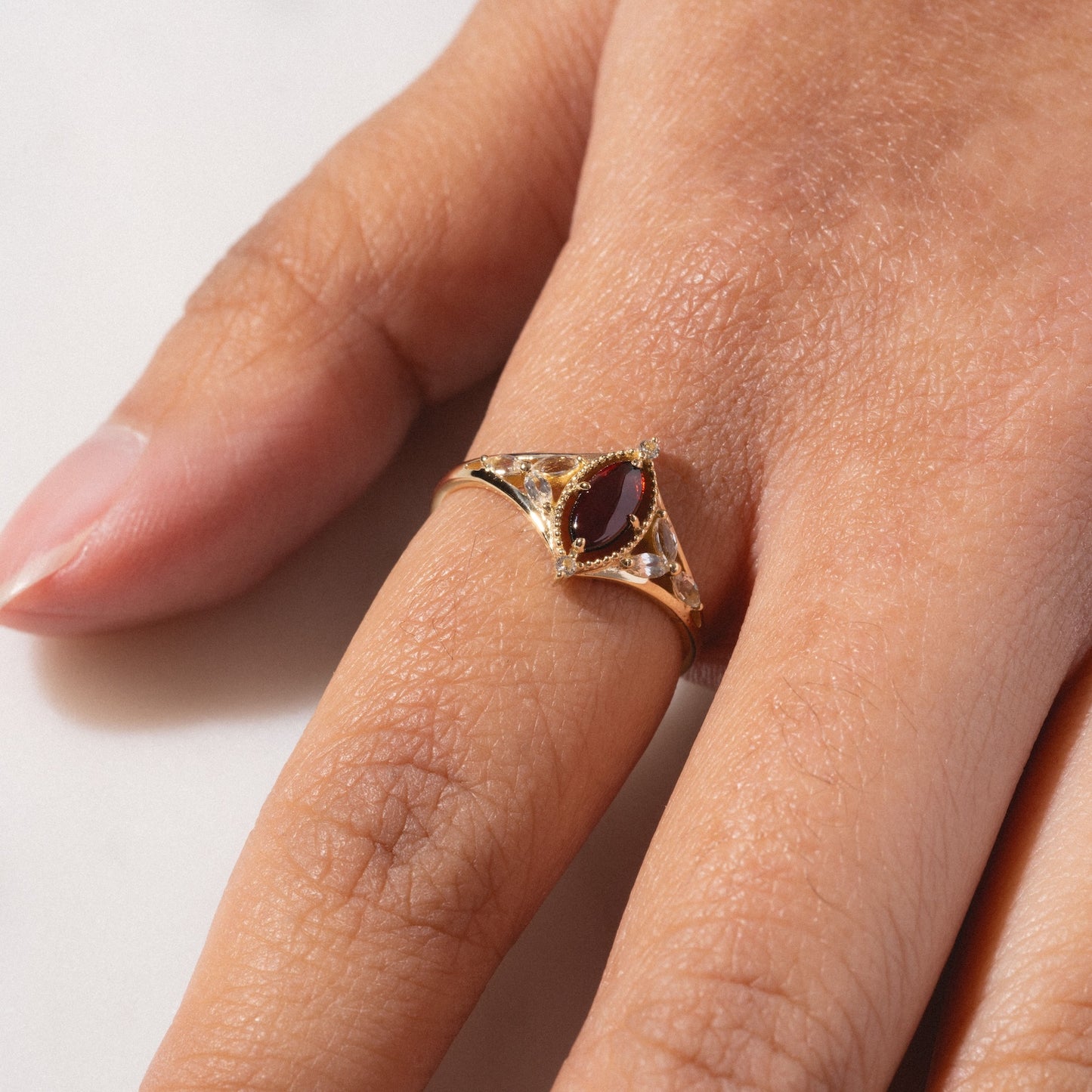 A close-up of a hand wearing the Solid Gold 2026 January Capsule Art Deco Garnet Ring, showcasing a marquise-shaped dark red gemstone with clear accents on the middle finger against a light background.
