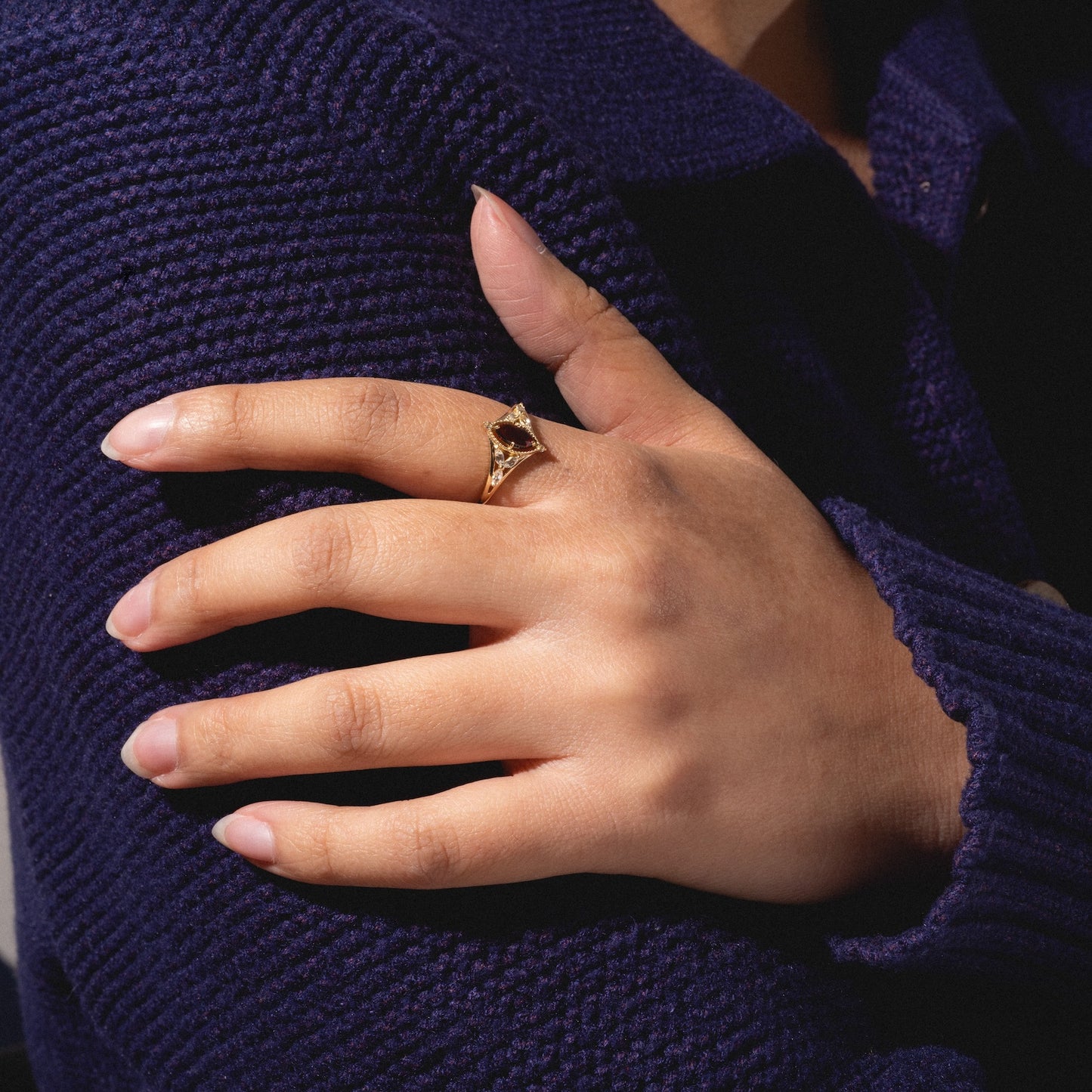 A person in a dark blue knitted sweater rests their hand on their arm, showcasing the Solid Gold 2026 January Capsule Art Deco Garnet Ring—the perfect January birthstone.