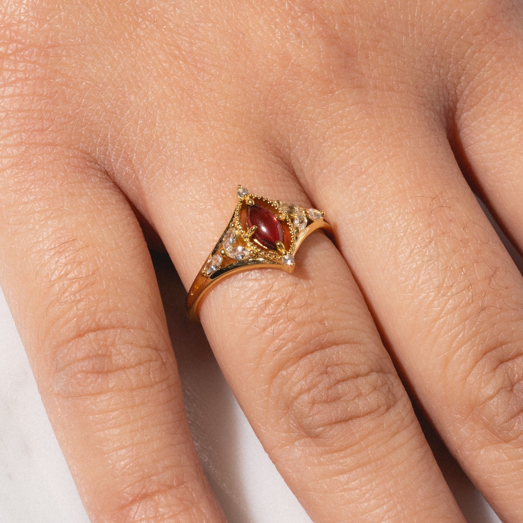 The 2026 January Capsule Art Deco Garnet Ring is a vintage-inspired yellow gold vermeil band with a red garnet and small clear stones, elegantly worn on the ring finger of a hand resting on a white surface.