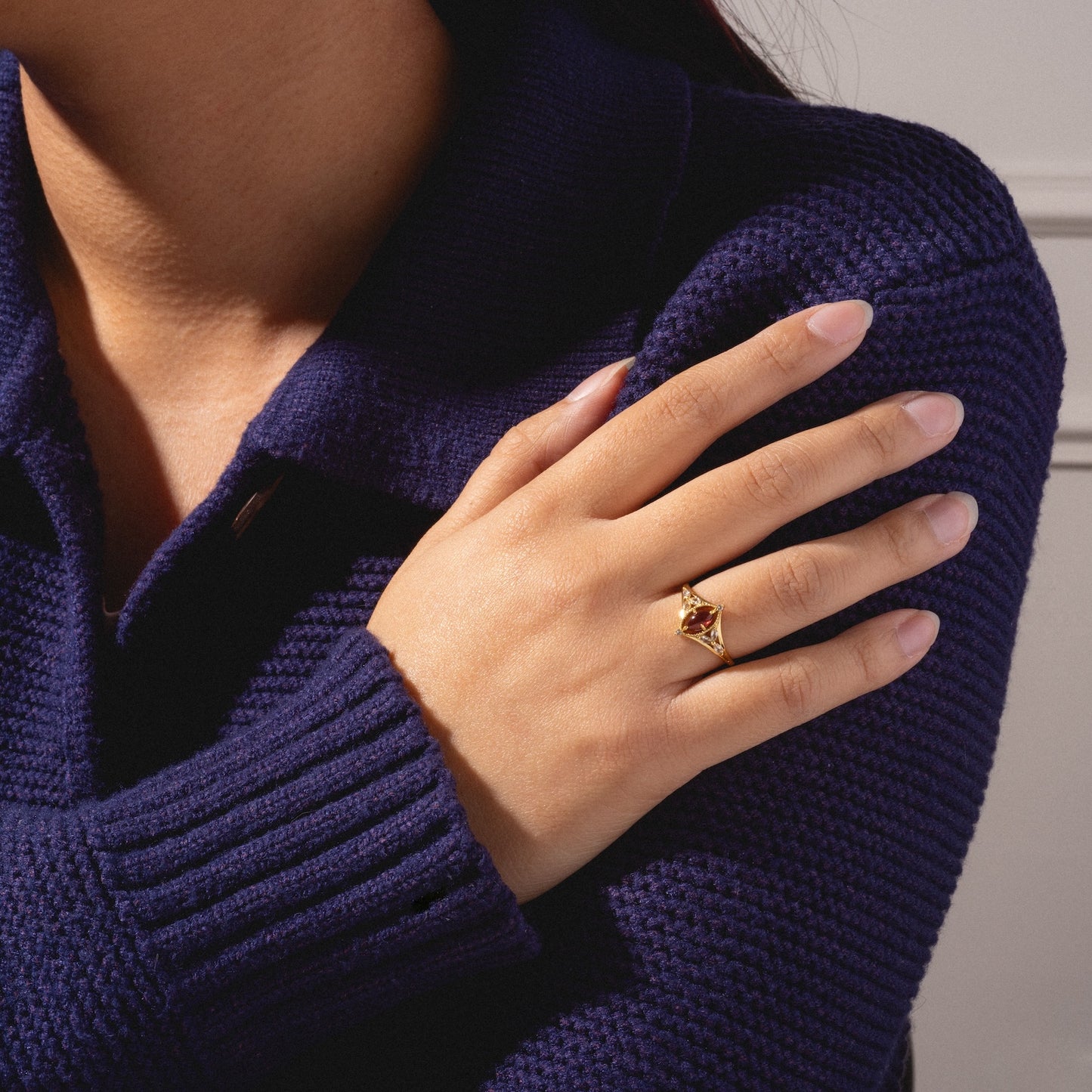 A person in a dark blue knit sweater crosses their arm, showcasing the 2026 January Capsule Art Deco Garnet Ring in yellow gold vermeil. The neutral background highlights the ring's vintage-inspired design and the textured sleeve.