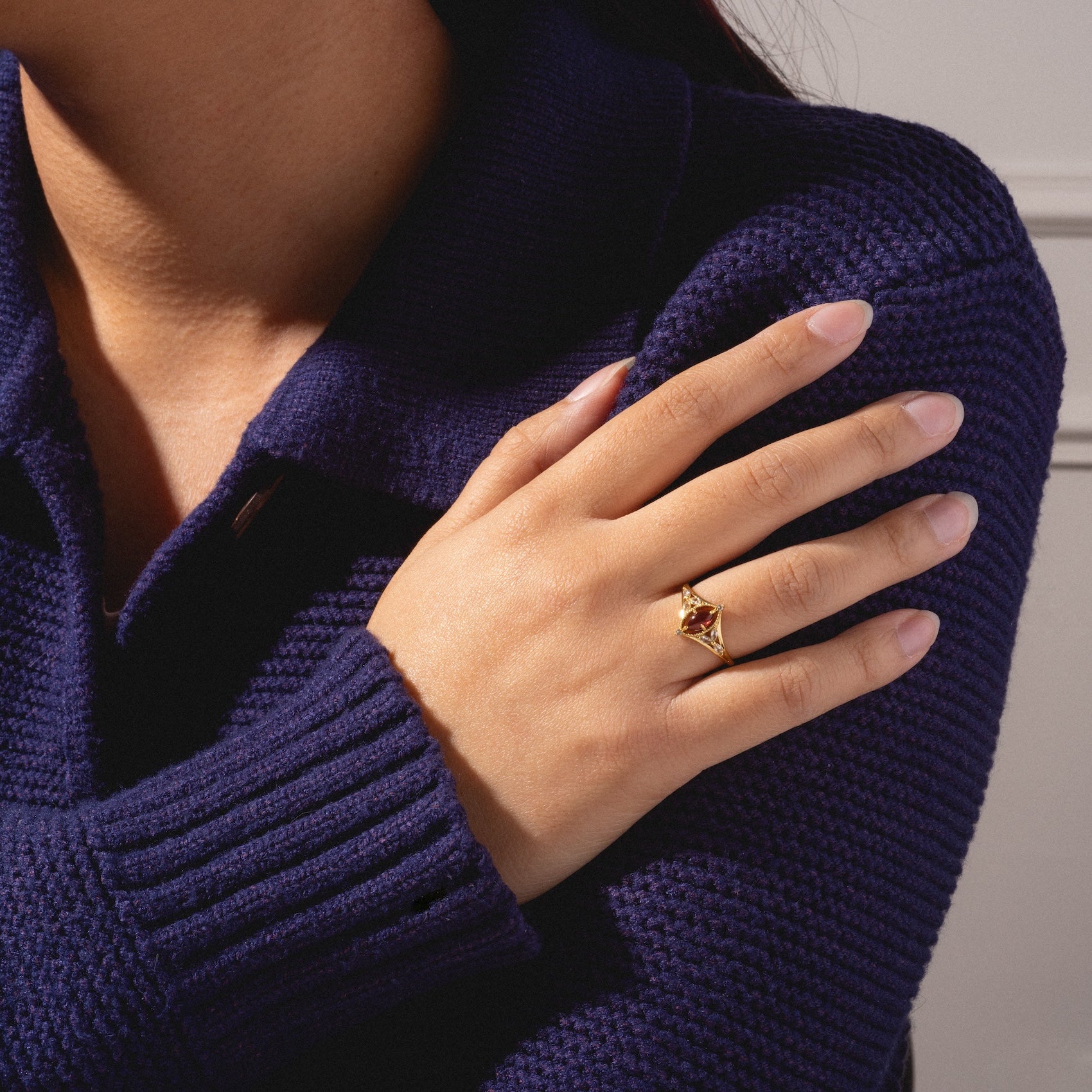 A person in a dark blue knit sweater crosses their arm, showcasing the 2026 January Capsule Art Deco Garnet Ring in yellow gold vermeil. The neutral background highlights the ring's vintage-inspired design and the textured sleeve.