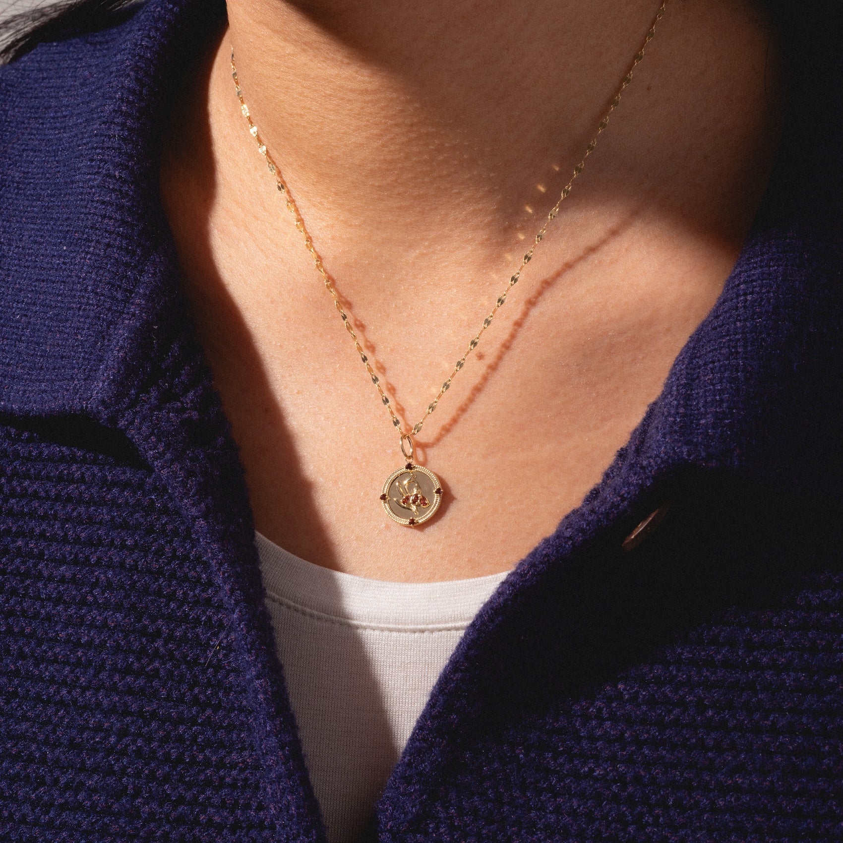 A person in a navy textured collared shirt and white top showcases the Solid Gold 2026 January Capsule Vintage Inspired Garnet Coin Charm, featuring a round pendant with a small engraved design.