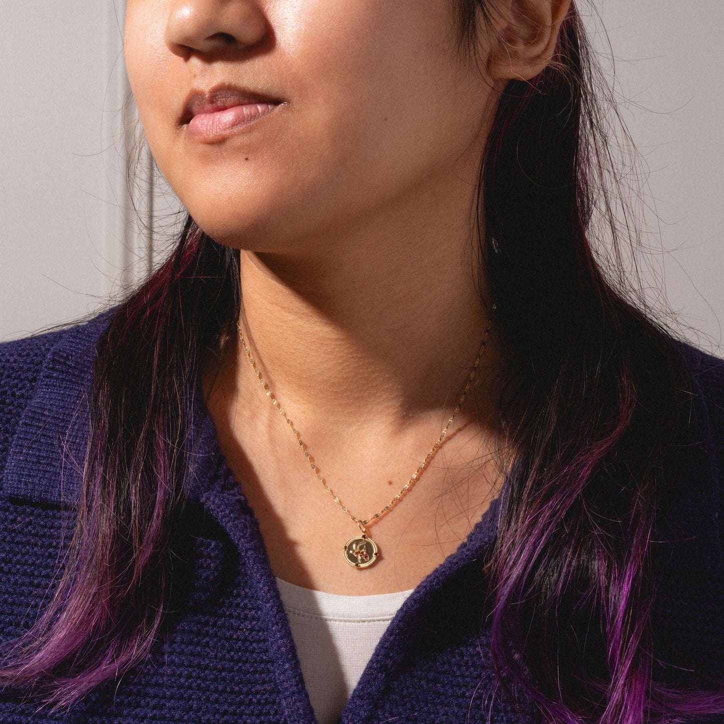 A woman with long dark hair and purple tips wears a blue textured jacket and the Solid Gold 2026 January Capsule Vintage Inspired Garnet Coin Charm necklace, standing before a plain light background. Only her lower face and upper torso are visible.