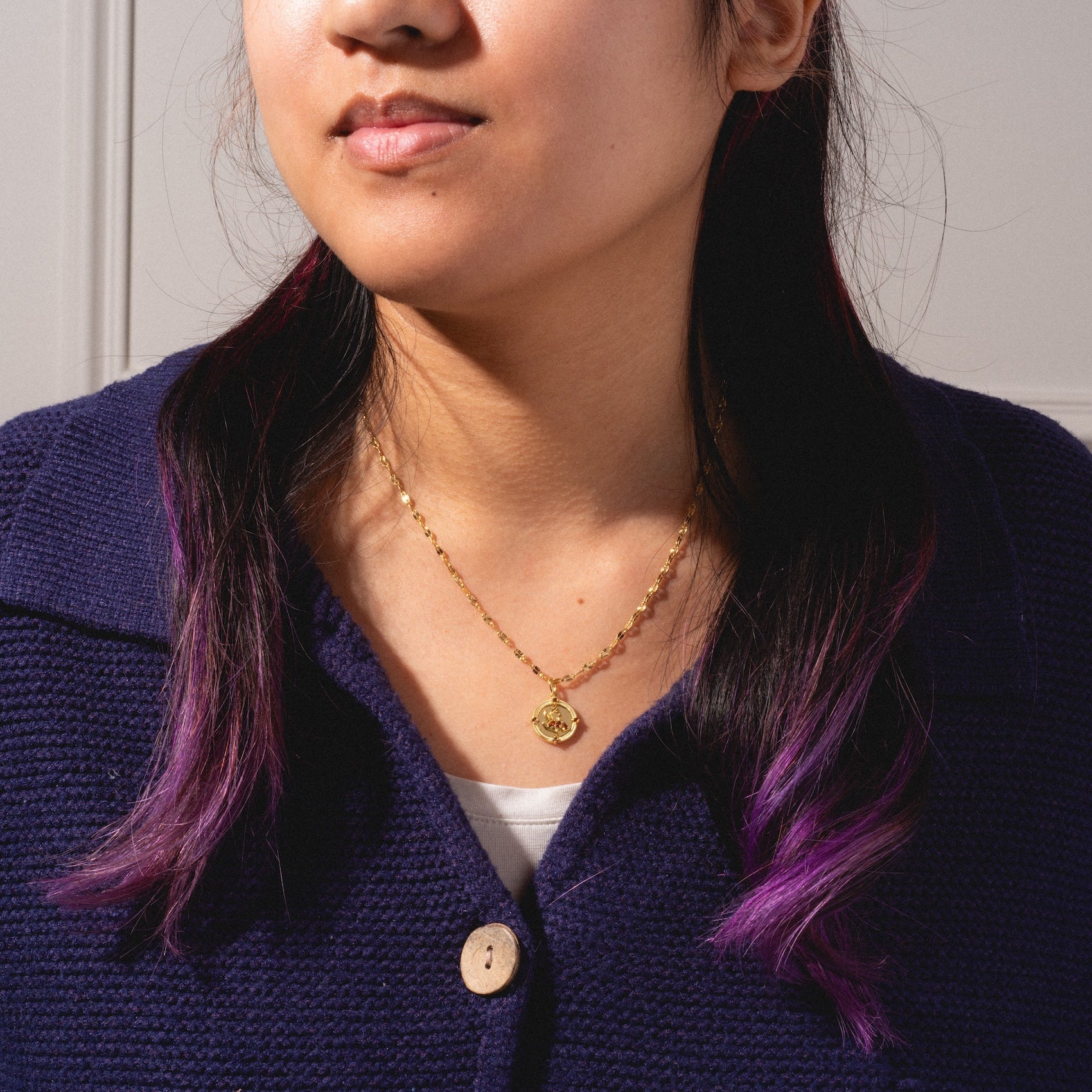 A woman with long dark hair and purple tips wears a navy blue textured cardigan and the 2026 January Capsule Vintage Inspired Garnet Coin Charm necklace, featuring a round pendant. She faces slightly to the side, showing only her lower face and upper torso.