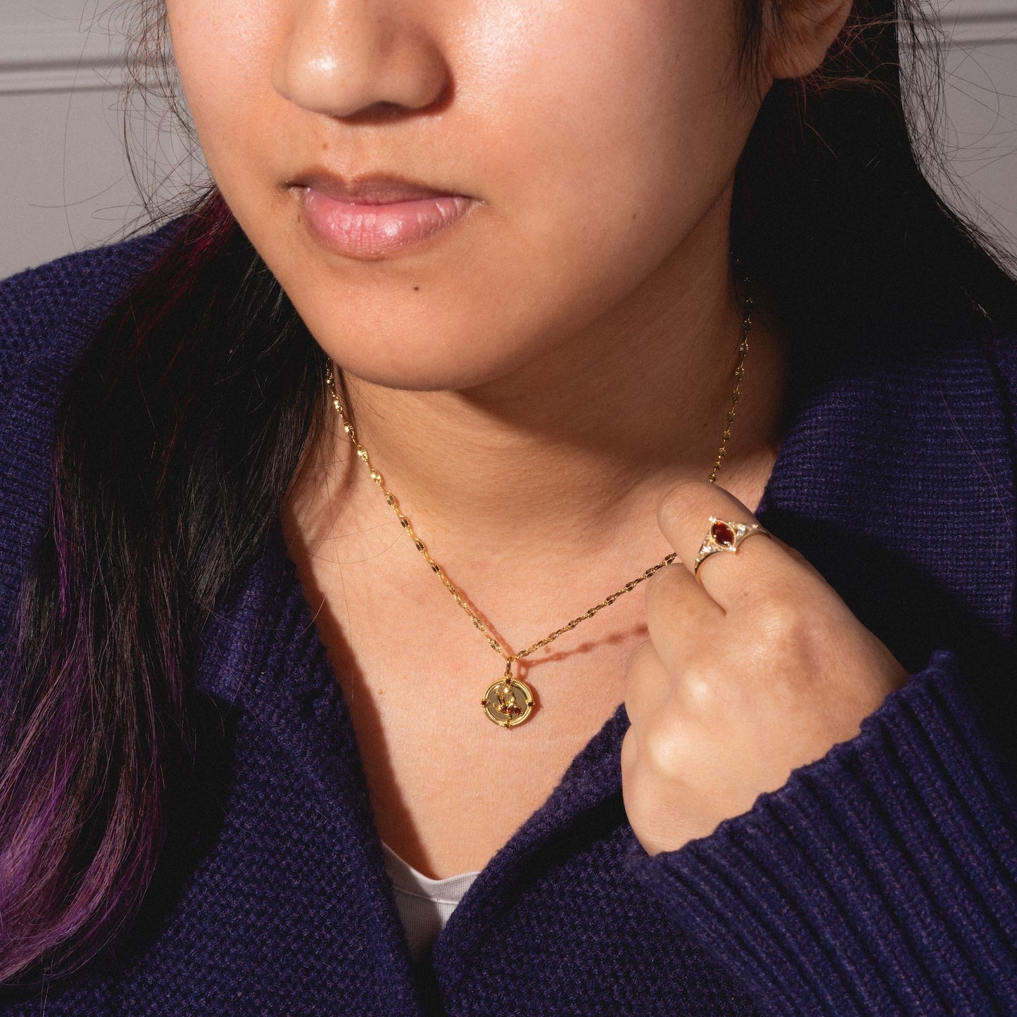 A woman in a navy sweater displays the 2026 January Garnet Capsule Set—with a gold round pendant necklace and a vintage-style gold ring set with a red garnet gemstone. Her hand grazes the necklace, and her hair shimmers with purple tones.
