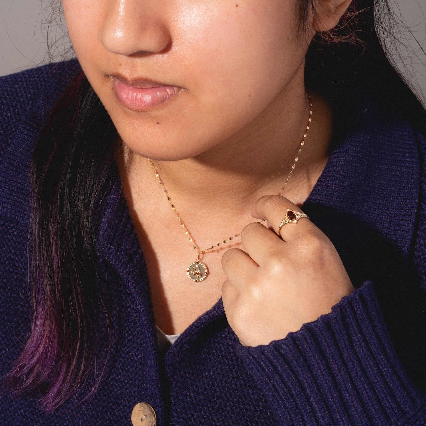 A woman in a navy blue sweater showcases the Solid Gold 2026 January Garnet Capsule Set, featuring a garnet pendant and a gold ring with dark gemstone. Her hair has a purple streak and her lips are closed. Free gift included.