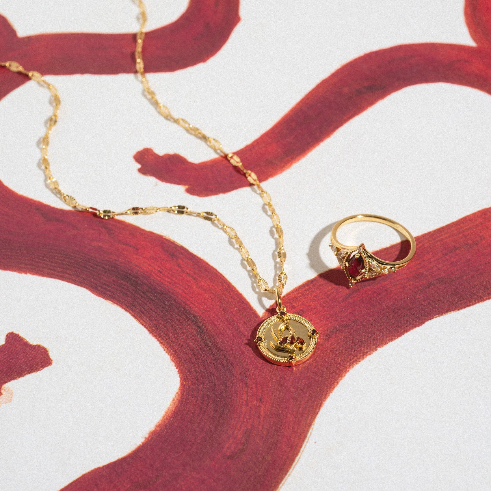 The 2026 January Garnet Capsule Set with Free Gift includes a vintage-inspired gold necklace with a circular garnet pendant and a gold ring with a red January birthstone, displayed on bold red abstract patterns.