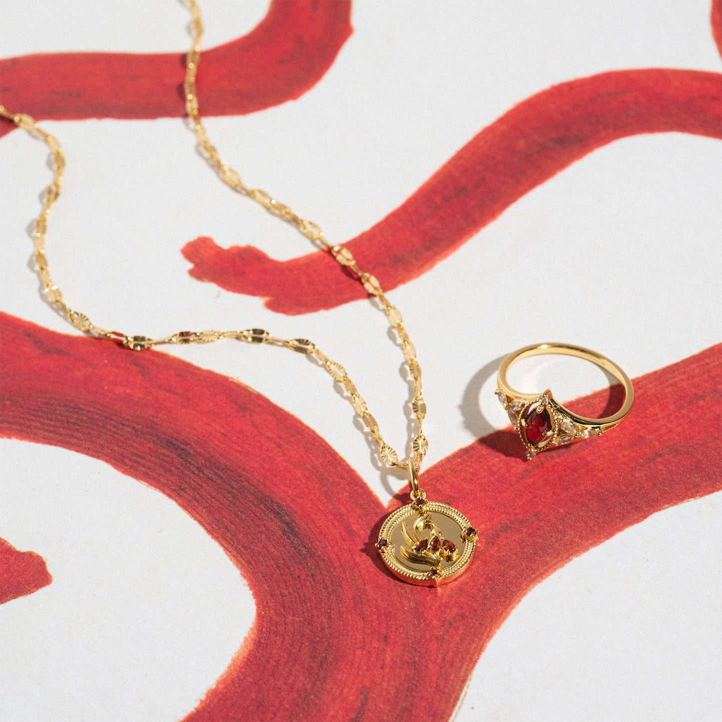 The 2026 January Capsule Art Deco Garnet Ring in yellow gold vermeil rests on a white surface painted with bold, curving red lines alongside a gold necklace with a round pendant.