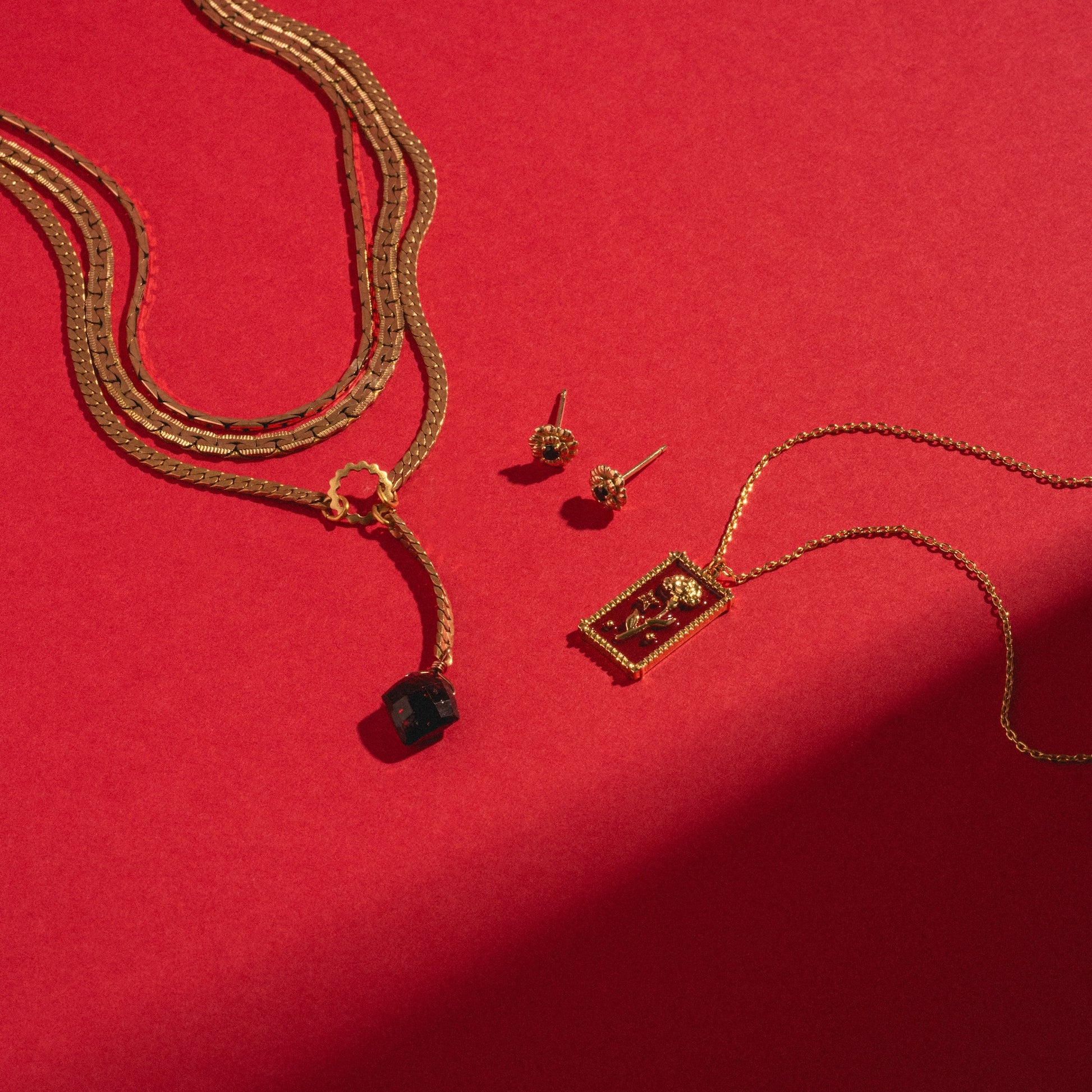 Gold necklaces and earrings are arranged on a red background. The Palmer Gemstone Necklace features recycled brass chains and a natural garnet pendant; another necklace has a rectangular charm, while the earrings have an intricate round design.