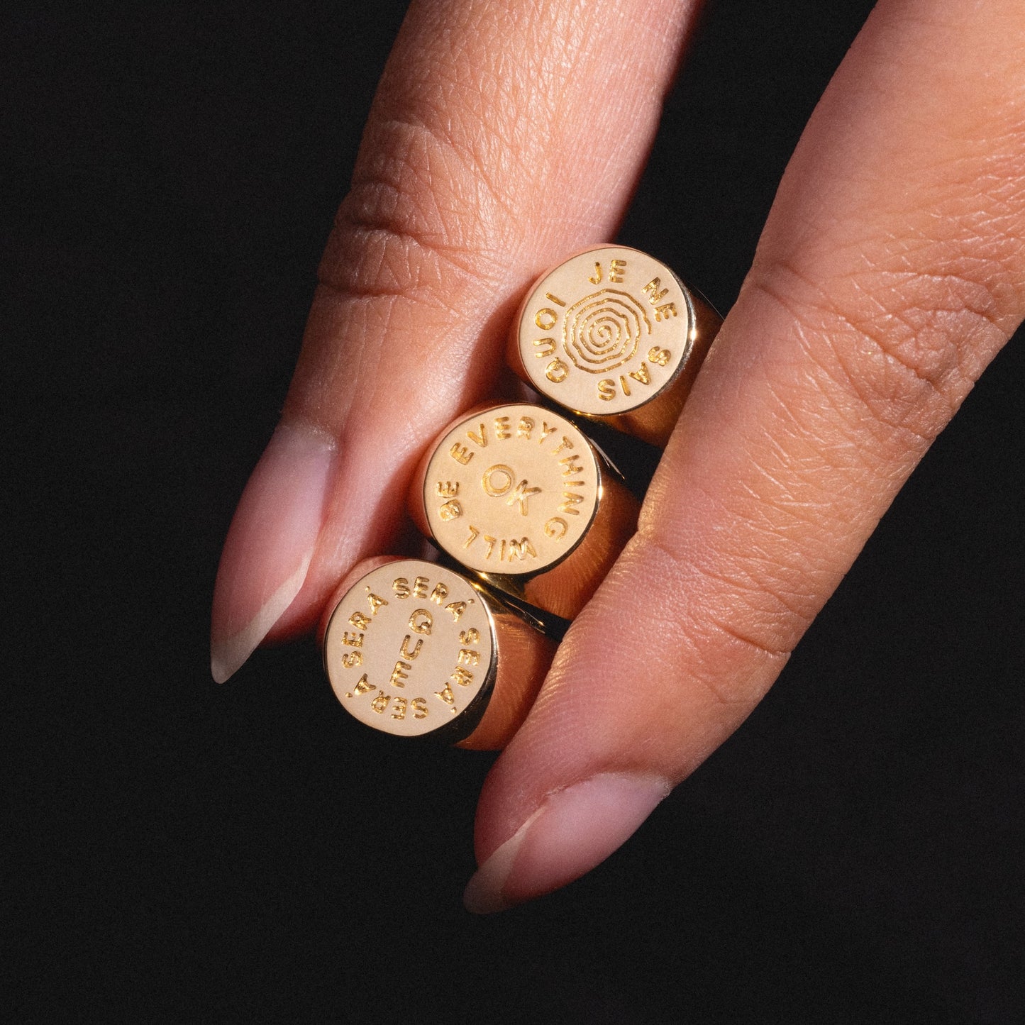 A hand with long, manicured nails holds three gold rings. One is the I Like It Here Club x Local Eclectic Je Ne Sais Quoi Pinky Ring, engraved with its namesake phrase. The other two read EVERYTHING WILL BE OK and SEA SEA SEA. Black background.