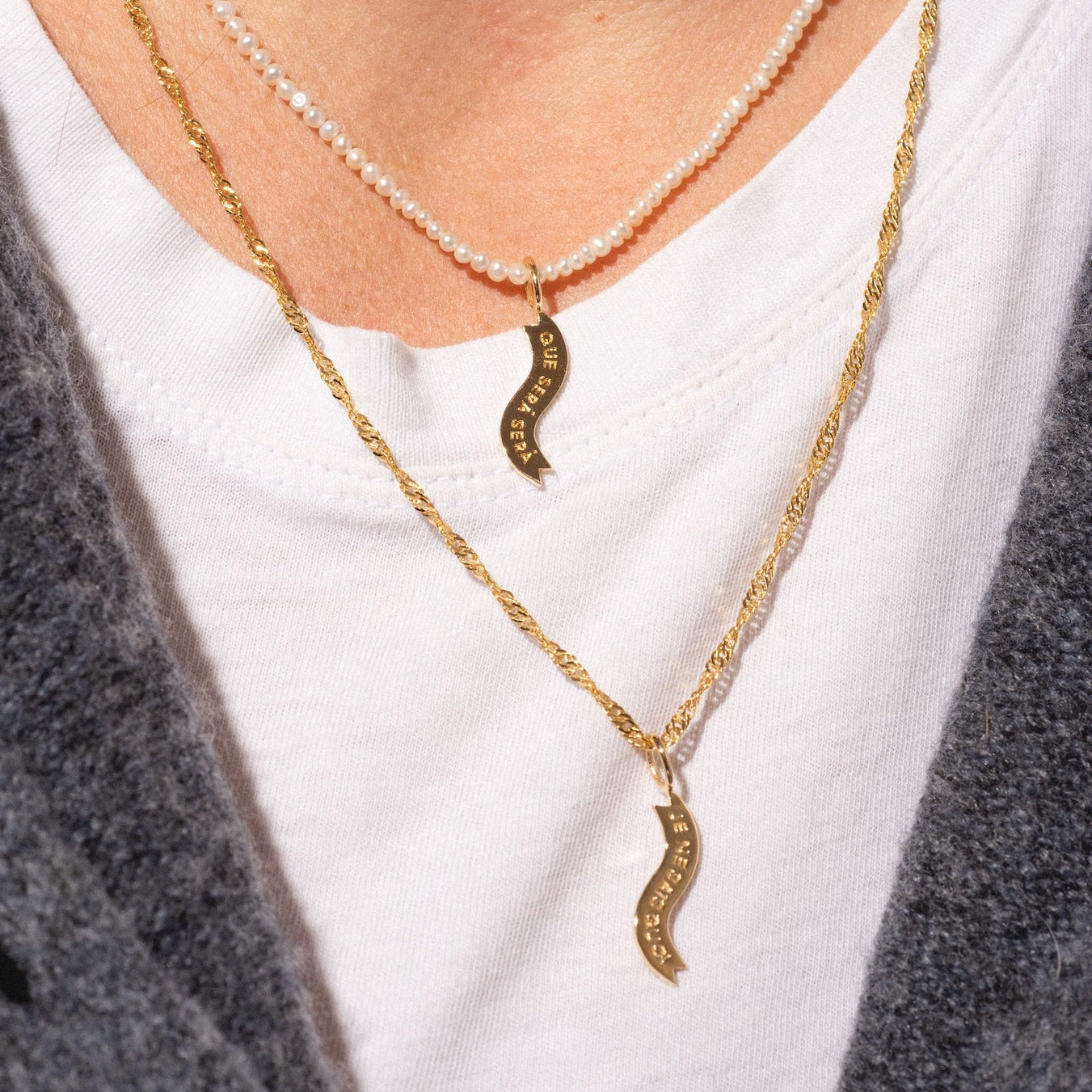 A person wearing two Charms by I Like It Here Club gold necklaces with matching wave-shaped charms, styled over a white crew neck T-shirt and a textured dark gray jacket.