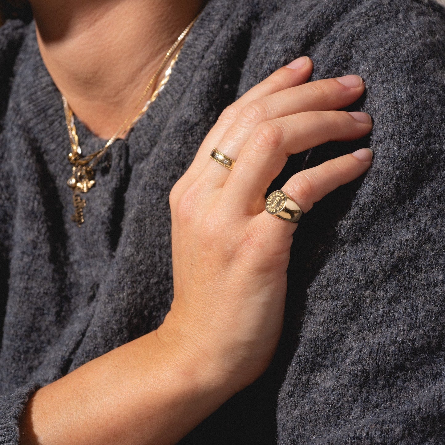 A close-up of a person wearing a dark gray sweater and layered gold necklaces, with their hand—adorned by the I Like It Here Club x Local Eclectic Que Sera Sera Pinky Ring—resting on their shoulder as sunlight casts gentle shadows.