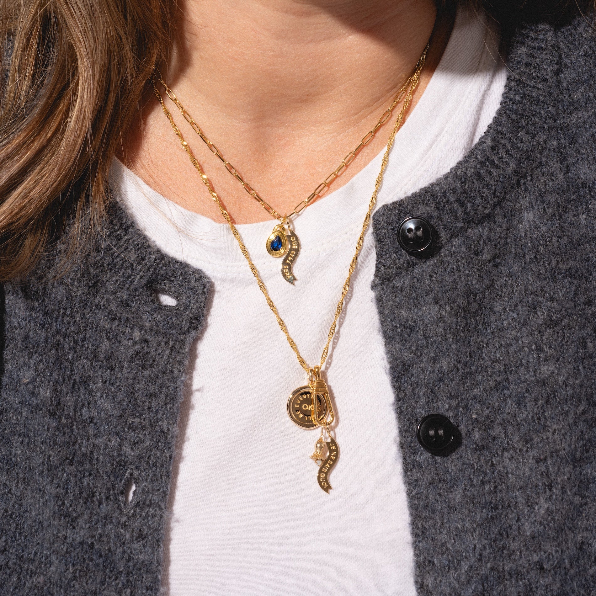 A person wears Charms by I Like It Here Club—layered gold necklaces with whimsical pendants like a blue gemstone, a circular gold plated charm, and curved horn-shaped charms—over a white shirt and dark gray textured cardigan.