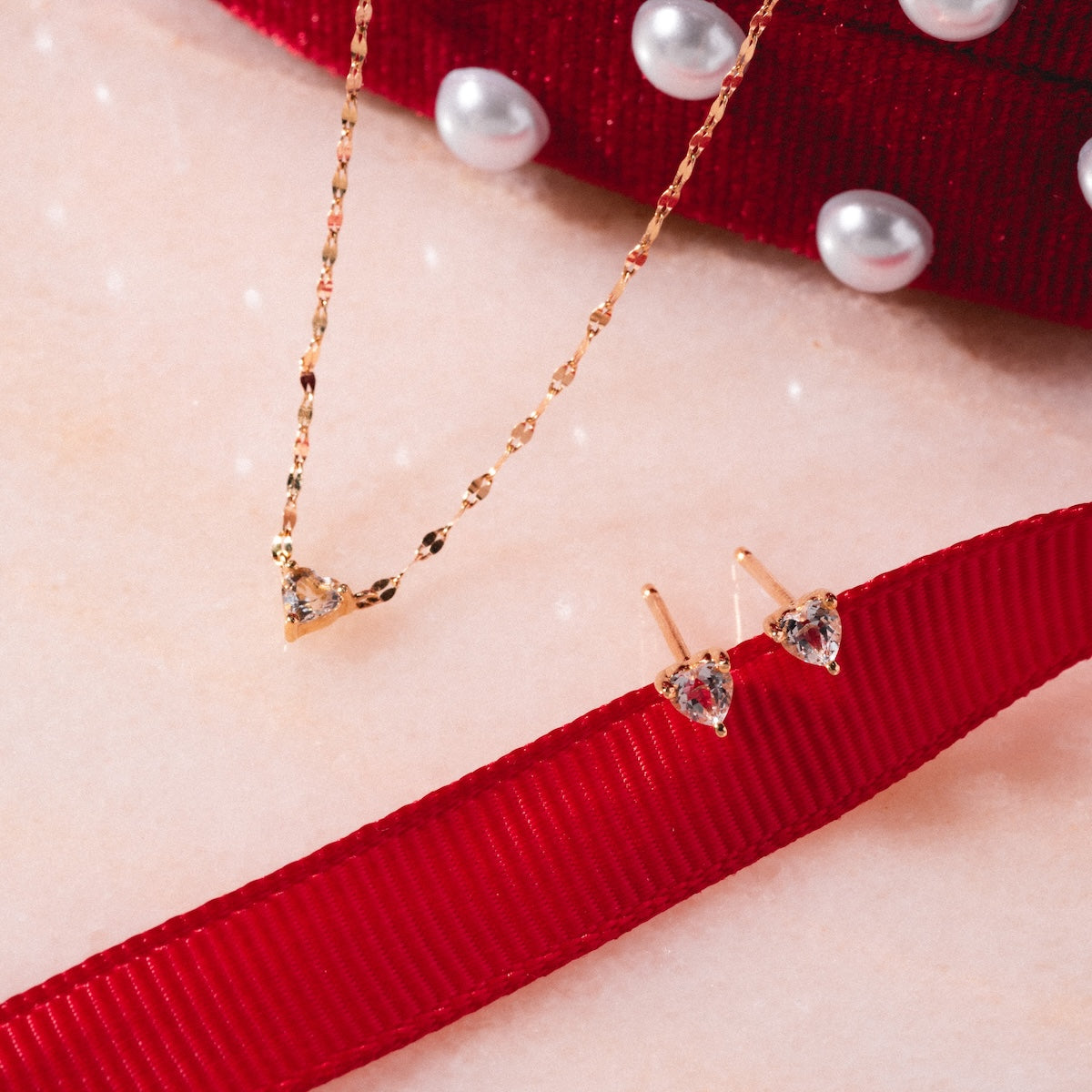 The Limited Edition Valentine’s Day Solid Gold Hearts Gift Set, featuring a heart-shaped pendant necklace and matching stud earrings with clear stones, is displayed on a light surface with a red ribbon and pearl-studded fabric in the background.