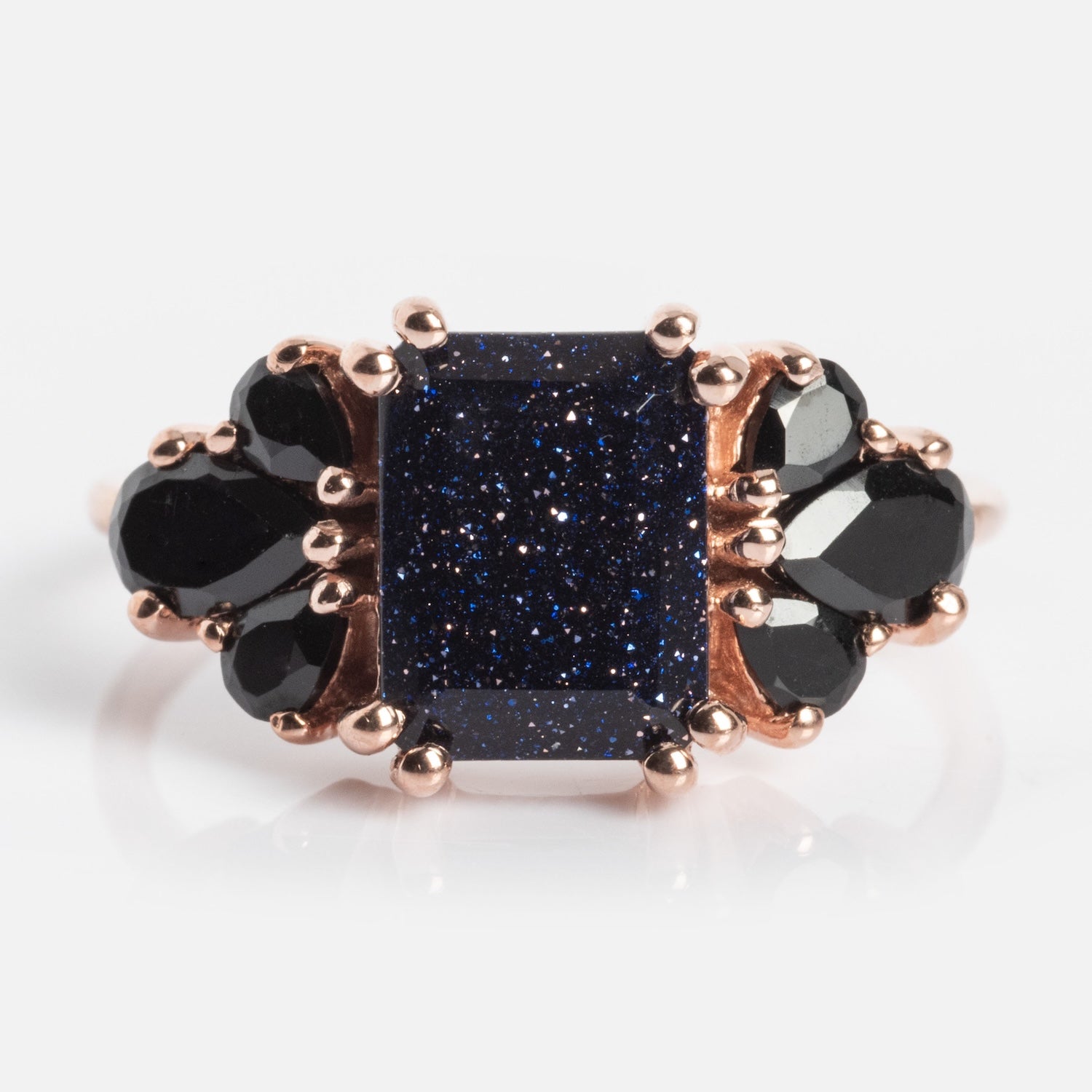 The Gemstone Empress Ring is a rose gold statement piece with a large rectangular blue goldstone center, sparkling surface, and clusters of faceted black gemstones on each side.