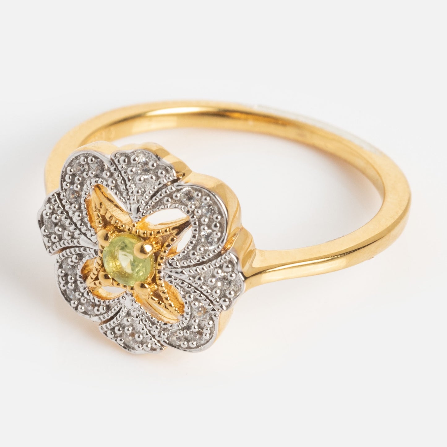 The Floral Art Deco Inspired Peridot Ring features a gold band with an ornate floral-shaped silver setting and a small green peridot gemstone at its center, shown against a white background.