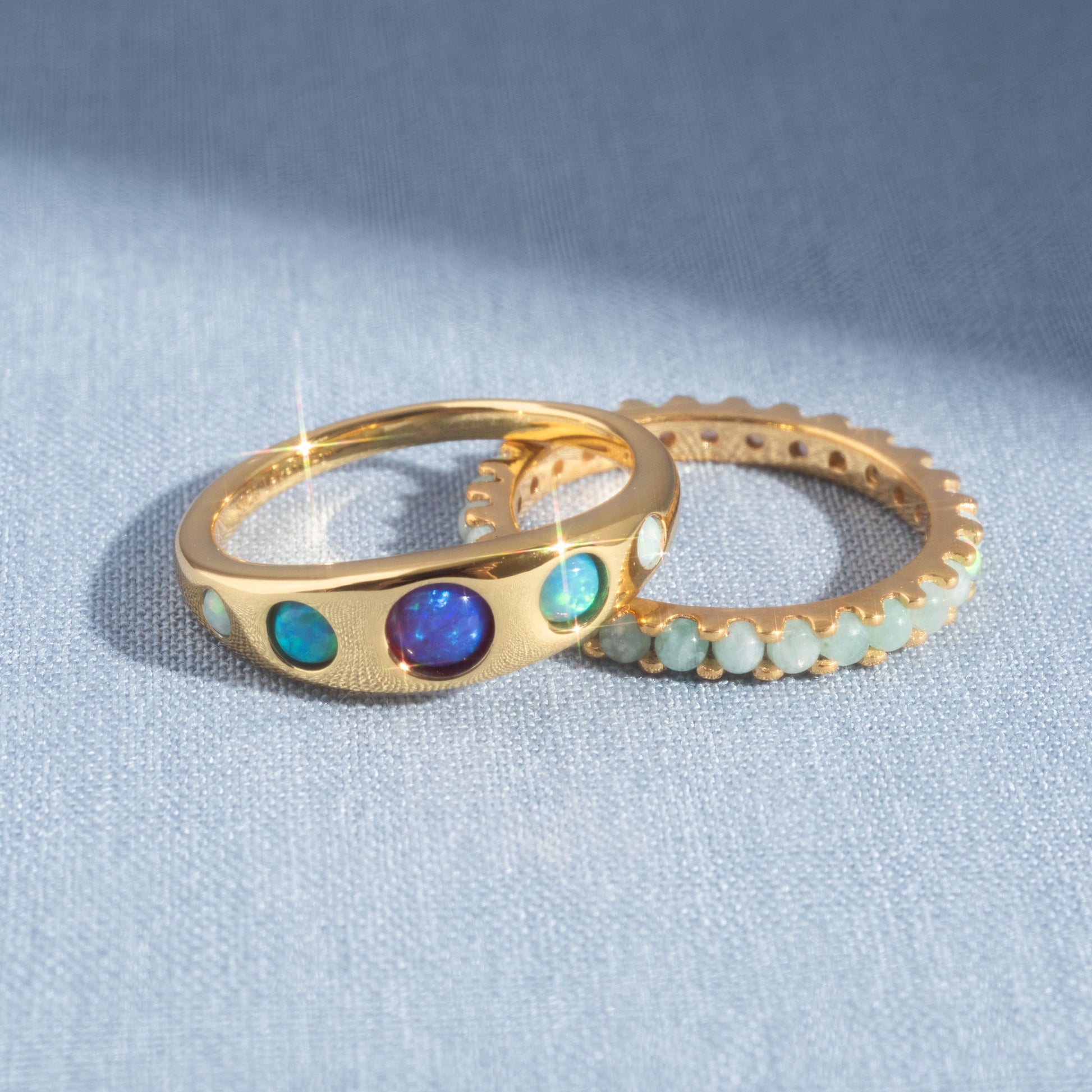 Two gold vermeil rings rest on a light blue textured surface: the Blue Opal Ombre Via Ring with iridescent stones, and another ring adorned with small, pale green beads encircling its band.