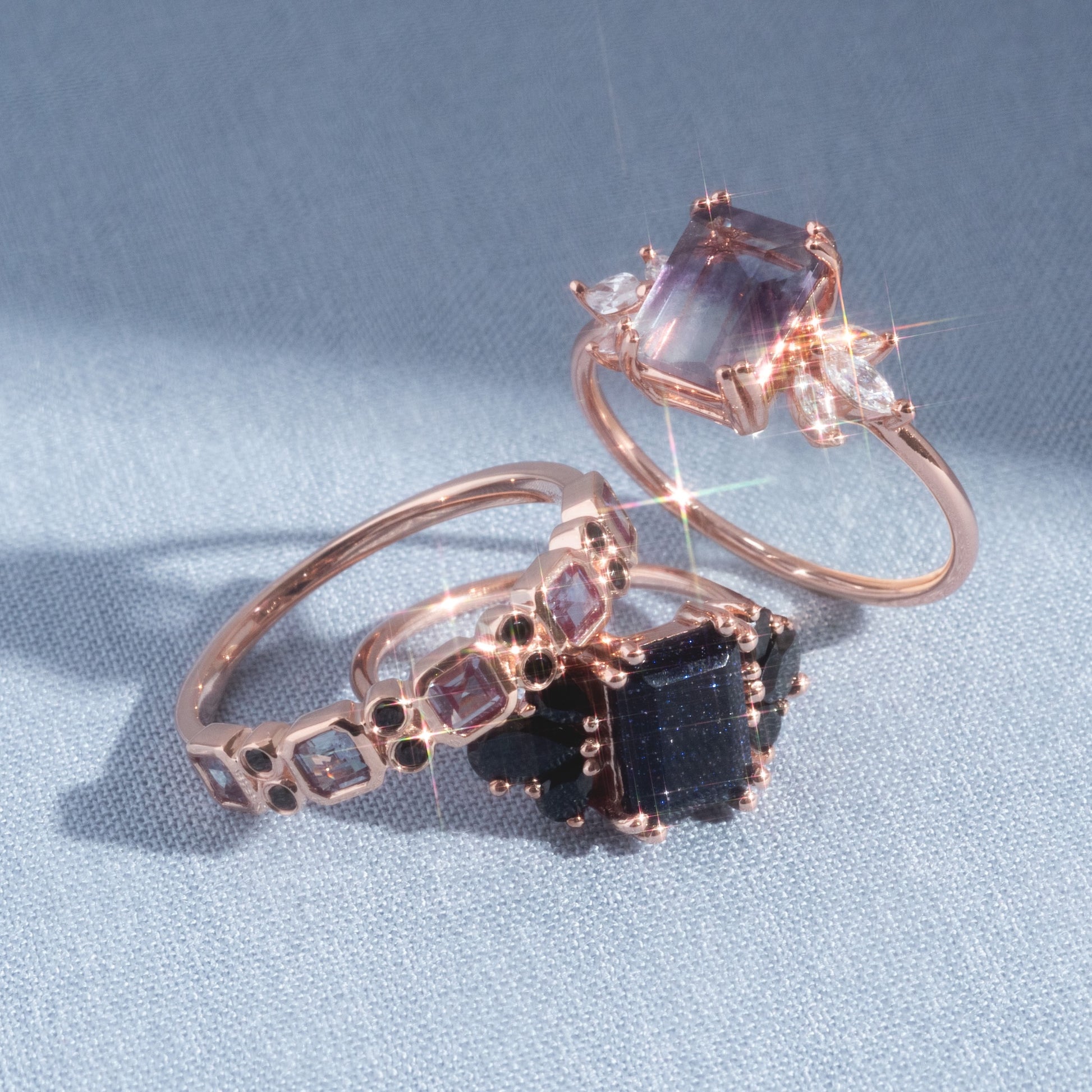 A 9kt rose gold Alexandrite & Black Diamond Juniper Ring with a large rectangular alexandrite gemstone and sparkling black diamond accents rests on pale blue fabric, catching the light beautifully.