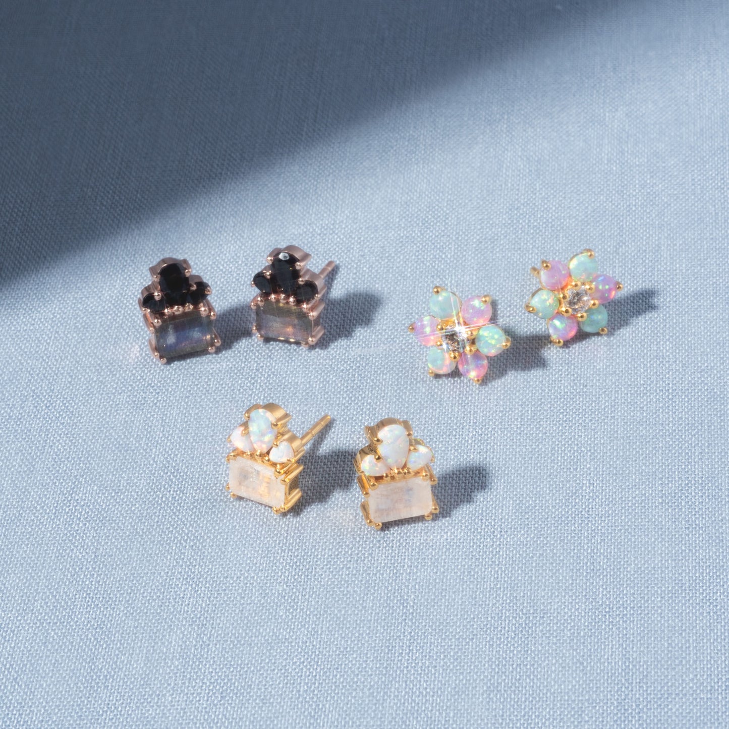 Three pairs of Gemstone Empress Stud Earrings are displayed on light blue fabric: one with black and clear stones, one with gold and iridescent pastel floral stones, and one in gold settings with square white stones.