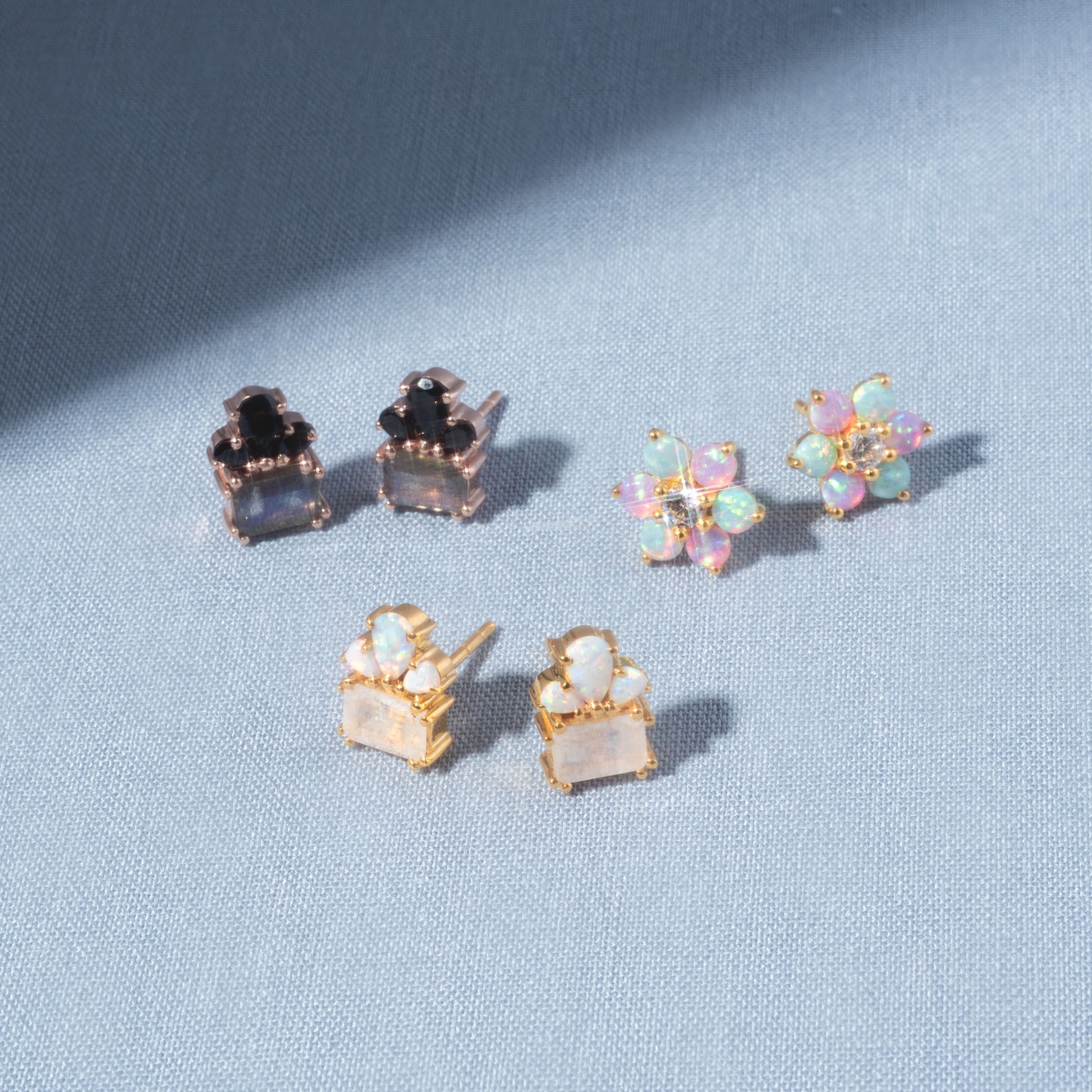 Three pairs of Gemstone Empress Stud Earrings are displayed on light blue fabric: one with black and clear stones, one with gold and iridescent pastel floral stones, and one in gold settings with square white stones.