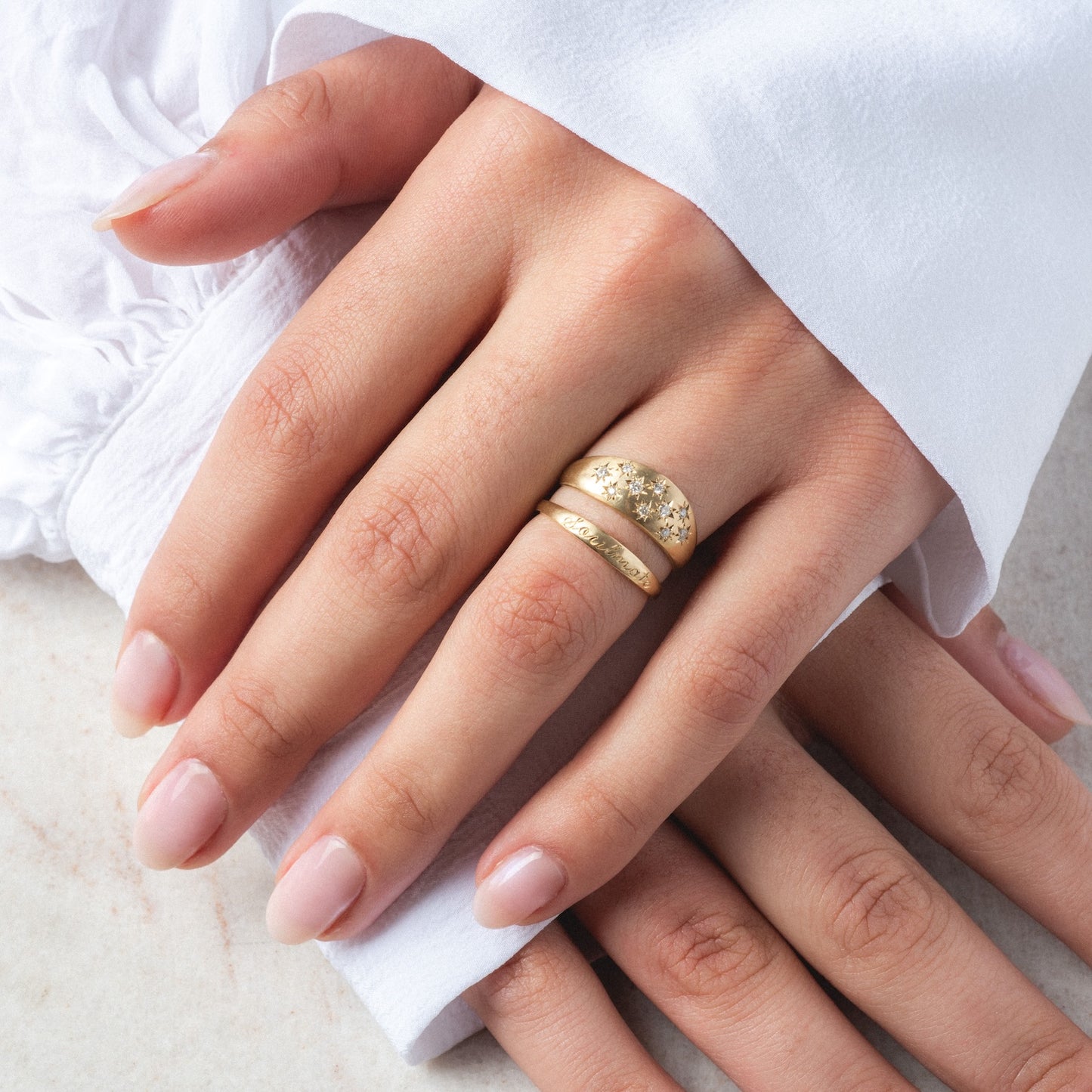 A hand with manicured nails rests on white fabric, wearing the Half Moon Bombe Ring alongside a textured gold ring and a delicate diamond band (0.22 ctw) on the middle finger. The person is dressed in a white long-sleeve garment.