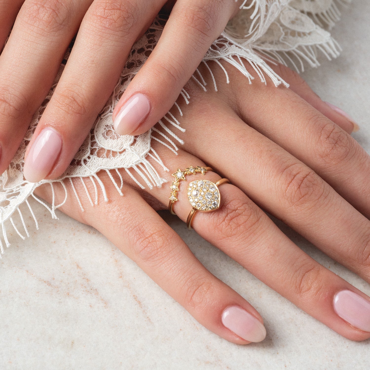 A close-up of hands with light pink manicured nails wearing gold rings, including the "My Beloved" Soul Ring diamond band, resting on white fringed lace fabric.