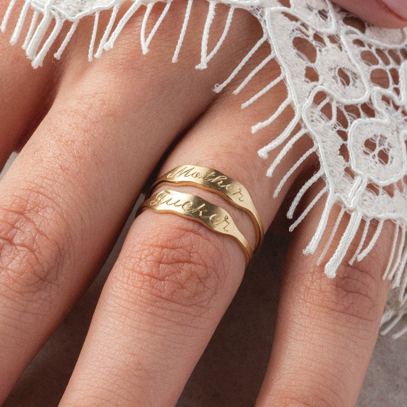 A close-up of a hand with light pink manicured nails, wearing two 14k Ripples Customizable ID Band rings, partially covered by delicate white lace fabric.