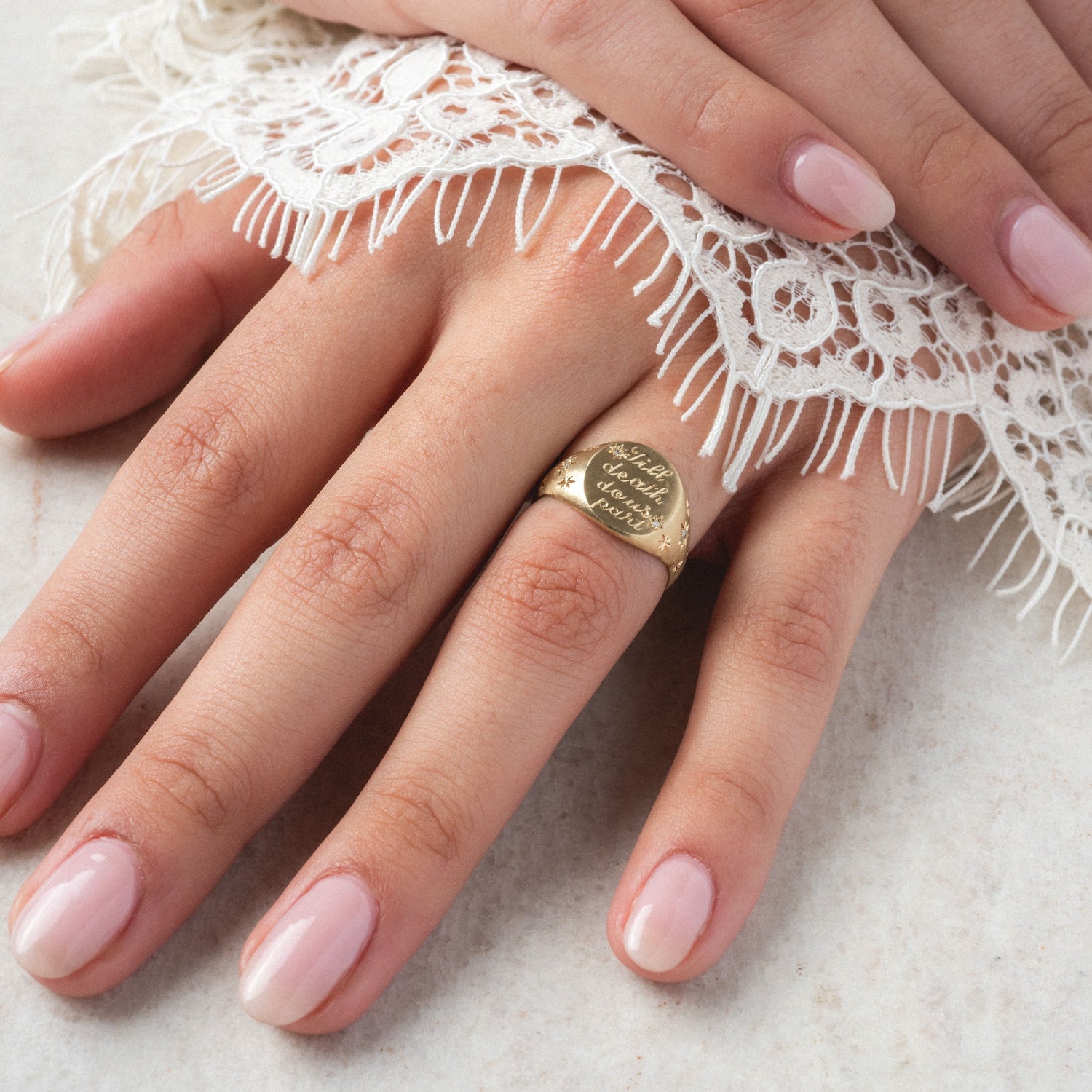 A hand with pale pink manicured nails rests on lace, showcasing the Till Death Do Us Part Signet with Diamonds—a gold ring on the ring finger engraved in cursive and accented with 0.04 ctw sparkling diamonds for an elegant touch.
