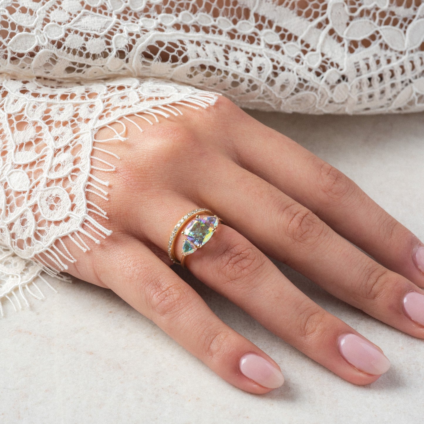 A manicured hand wears the Seraphina Ring—a gold band with a marquise-cut angel aura quartz center and small round stones—paired with a fringed white lace sleeve.