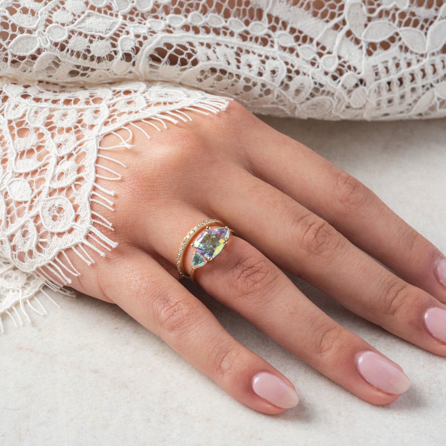 A manicured hand wears the Seraphina Ring—a gold band with a marquise-cut angel aura quartz center and small round stones—paired with a fringed white lace sleeve.