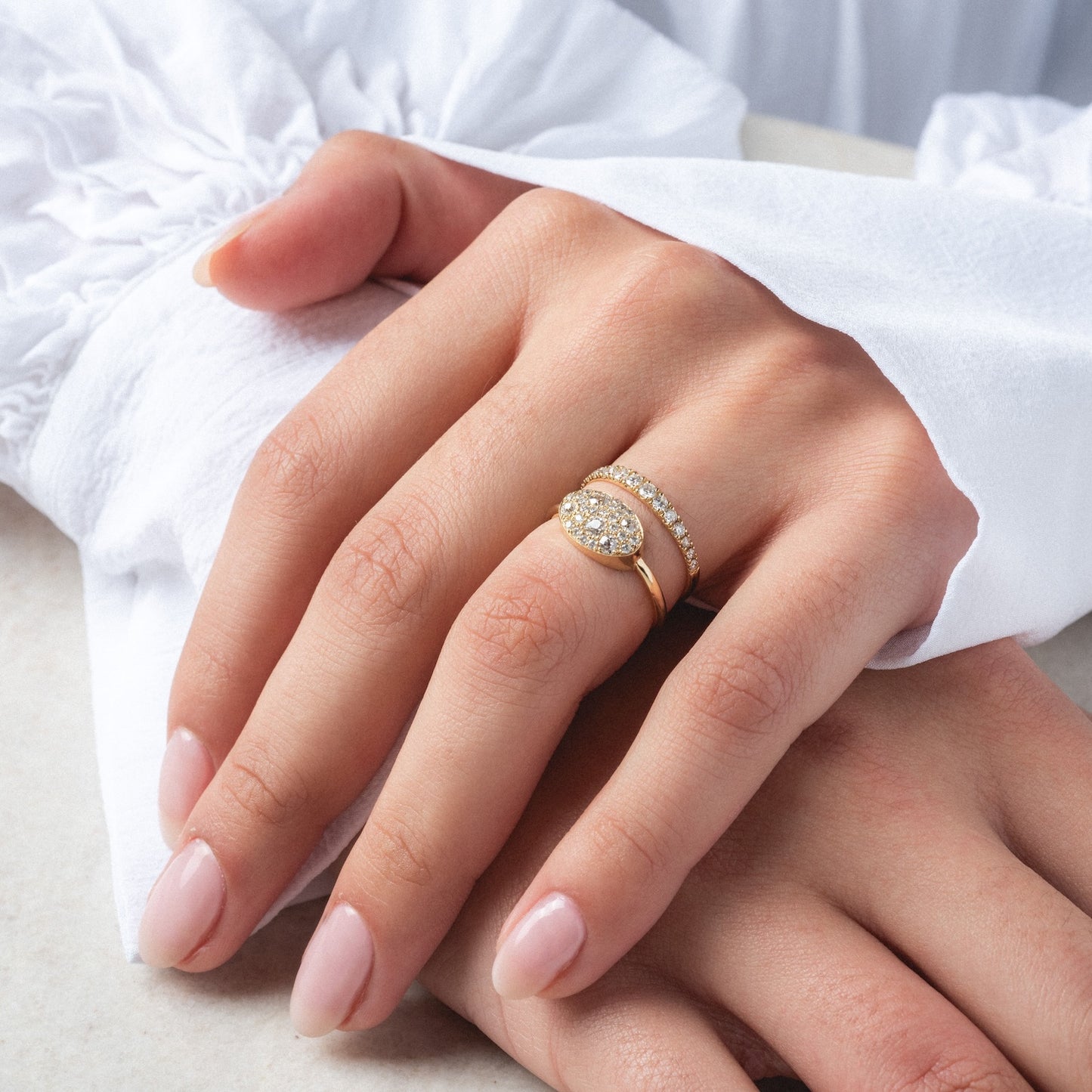 A hand with manicured nails rests on a white surface, wearing the "Eternal Flame" Soul Ring—a slim 1.5 mm gold band that sparkles with small diamonds beside an oval cluster of old mine cut diamonds on another ring.