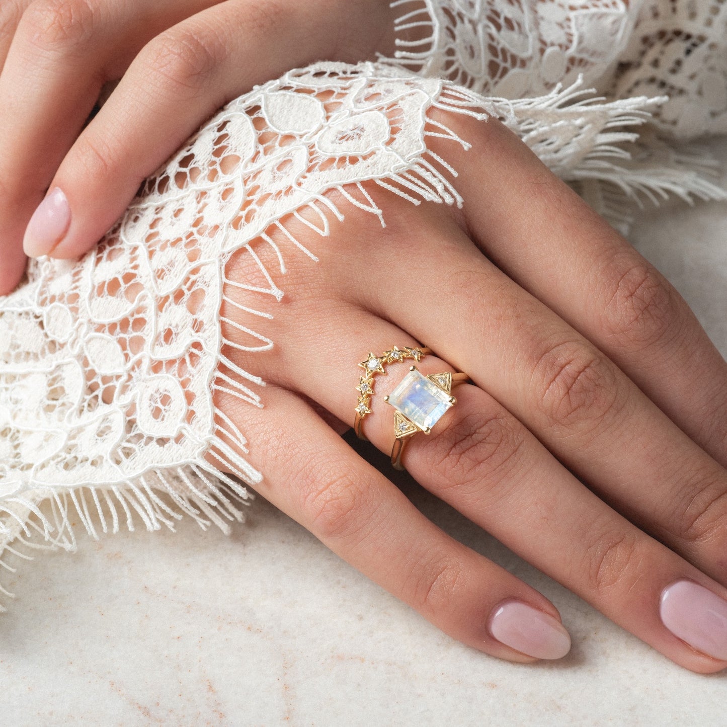 A hand with light pink manicured nails wears two gold rings—one is the Celestia Ring, featuring a large rectangular rainbow quartz stone and star accents. The sleeve is white lace with fringe, resting on a smooth surface.